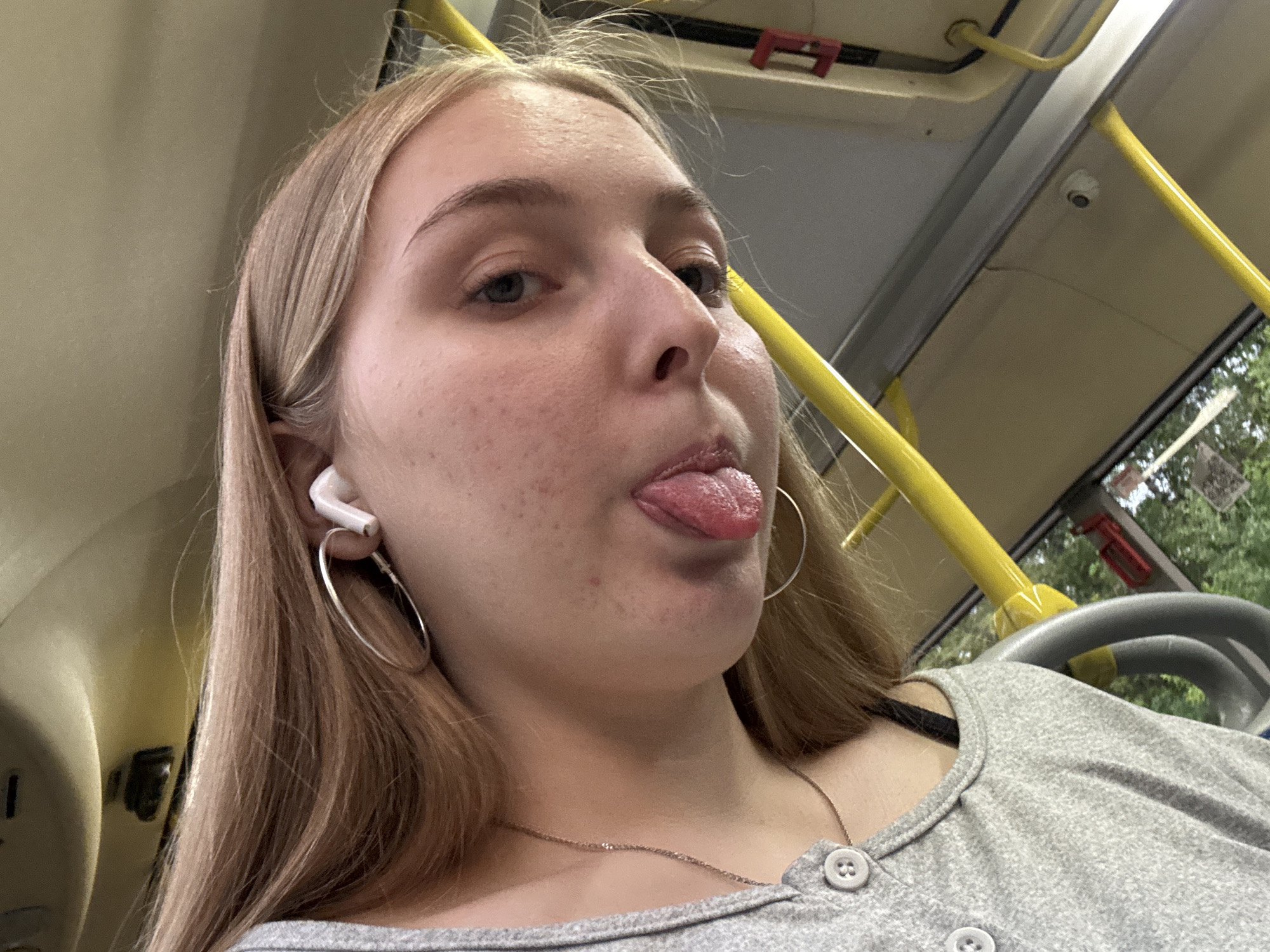 A young female with light brown hair, light eyes, and fair skin, exhibiting minor facial blemishes, is depicted in the interior of a public transport vehicle in the city of Perm, Russia. She is shown from a low-angle perspective, making a playful expression by extending her tongue. She wears a light grey short-sleeved top with three visible grey buttons down the front, a thin gold-colored chain necklace, a large silver hoop earring in her left ear, and a white wireless earbud in the same ear. The environment is characterized by prominent yellow grab rails and overhead handrails, beige interior panels, and a small white security camera mounted on the ceiling. Through a window on the right, green foliage and urban structures are partially visible.