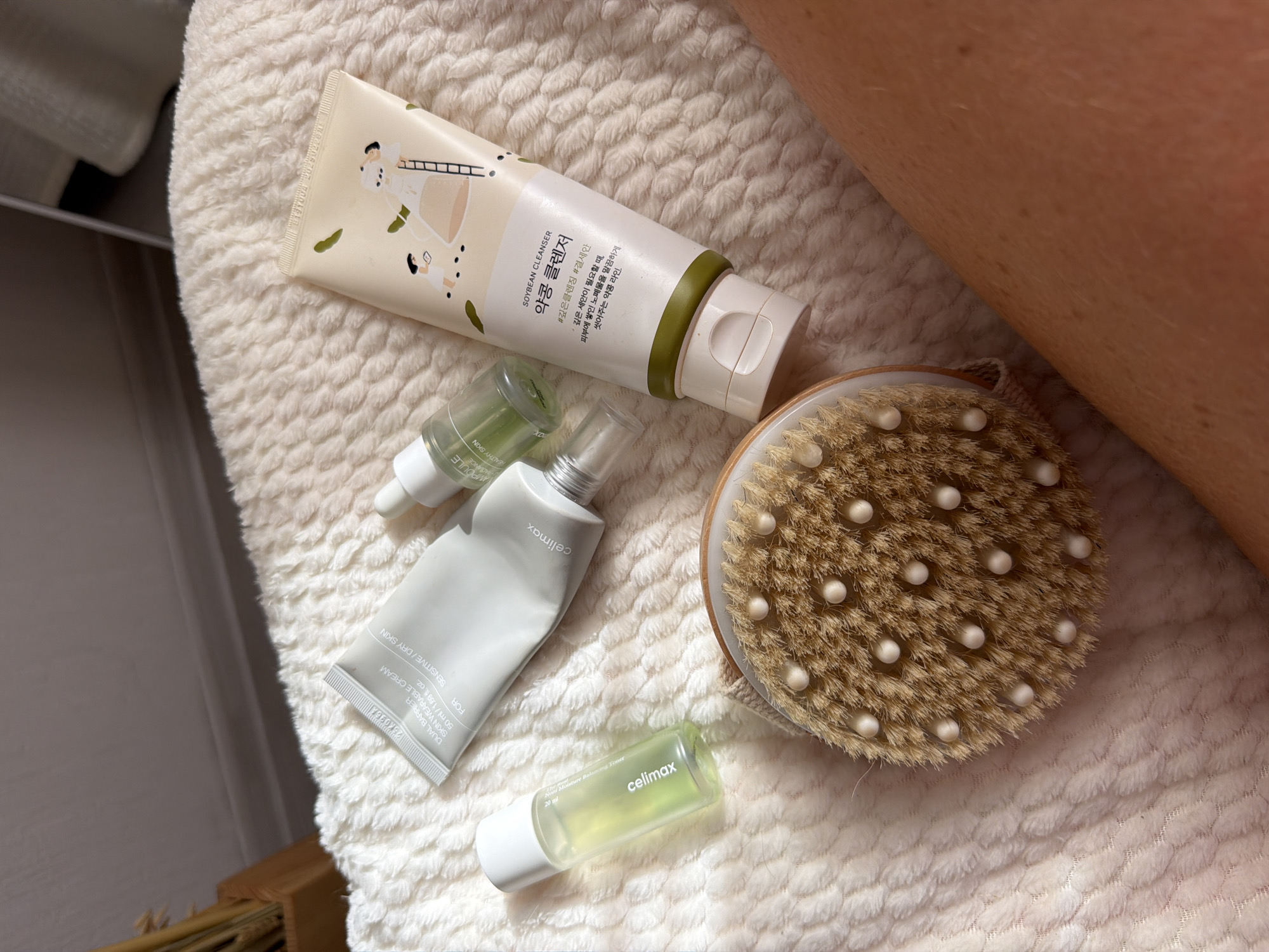 A high-angle, close-up shot shows an assortment of skincare products and a dry brush laid out on a textured, cream-colored blanket. The products include a gray tube labeled "DUAL BARRIER SKIN WEARABLE CREAM", a small clear bottle with green liquid and a dropper lid, and a larger cream-colored tube with an illustration of a person on a ladder and Korean text that reads "SOYBEAN CLEANSER". A small, clear bottle with green liquid is also visible. In the foreground, a round dry brush with a wooden handle and natural bristles, studded with small white balls, is positioned diagonally. The background is out of focus, showing a hint of a wooden object and a wall. The scene is illuminated by natural light, casting soft shadows. There are no visible people or indications of the city of Moscow, Russia.