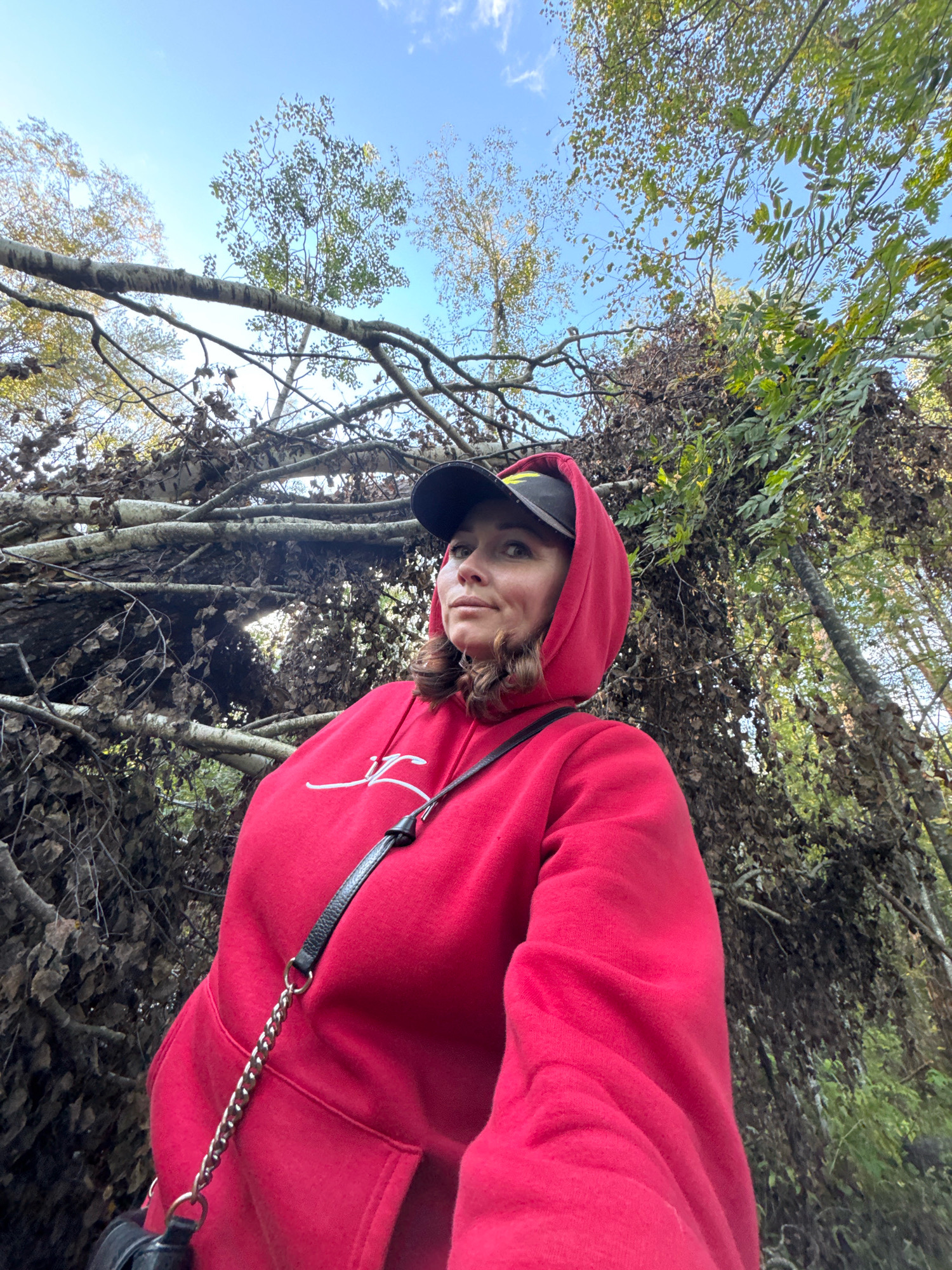 A woman is captured from a low angle, looking up and to the right, against a backdrop of trees and a bright blue sky. She is wearing a red hooded sweatshirt with a white graphic on the chest and a black baseball cap. A black purse with a silver chain strap is visible. The environment appears to be a wooded area with fallen branches and foliage. The location is identified as Innolovo, Russia.