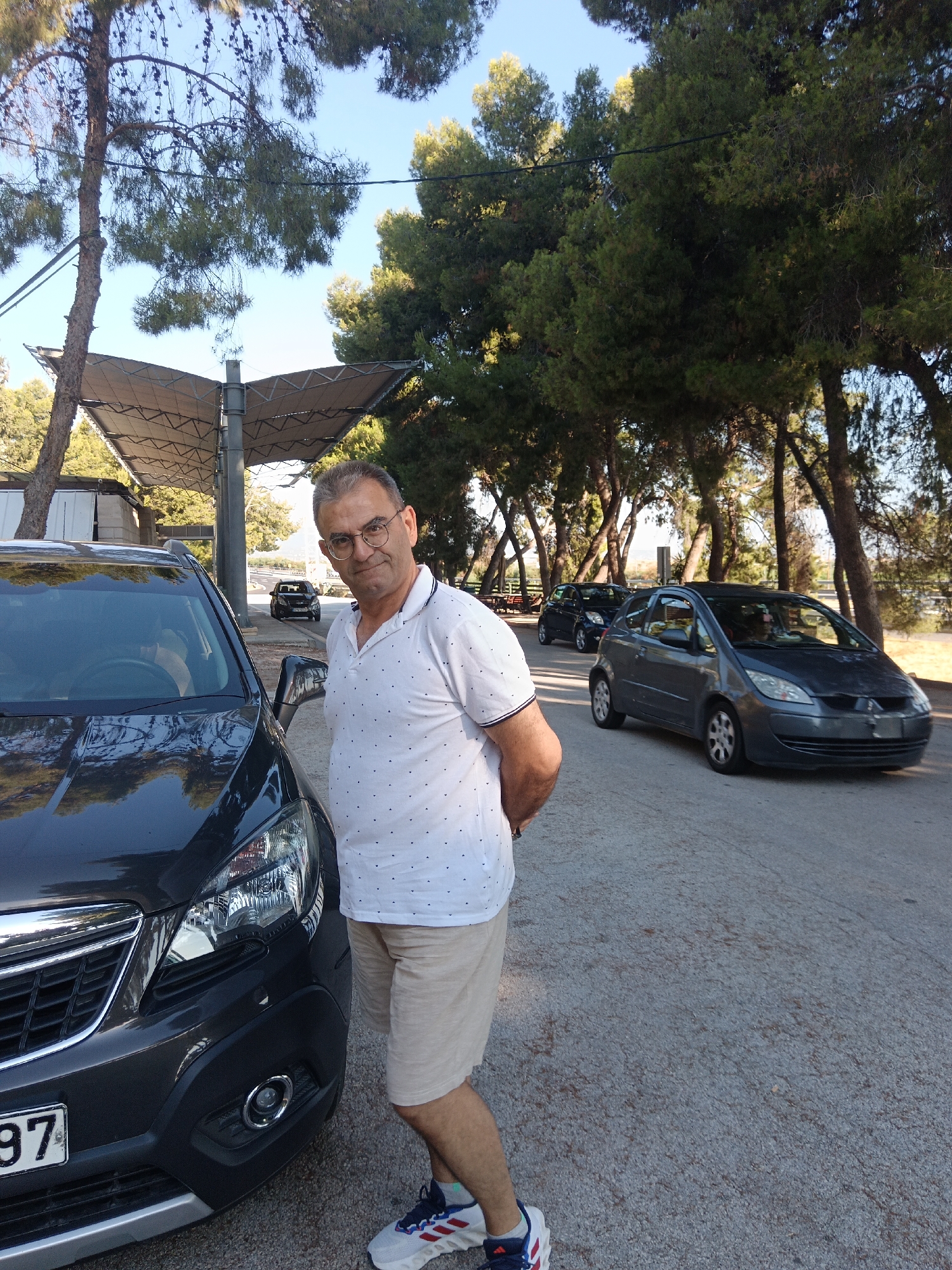 A man in his 50s stands next to a dark gray SUV, partially obscuring its front left side. He wears glasses, a white polo shirt with small dark blue polka dots, and tan shorts. He has his hands behind his back. The SUV has a license plate that reads "97". Behind the SUV, a dark gray hatchback is parked on the right side of a paved road. Further down the road, another dark hatchback is visible, as well as a covered outdoor seating area or pavilion. The area is lined with trees, primarily pine trees, casting shadows on the ground. The sky is clear and blue. The scene appears to be a parking area in a natural setting, possibly a park or a roadside rest stop. This location is within the Municipal Unit of Vocha, Greece.