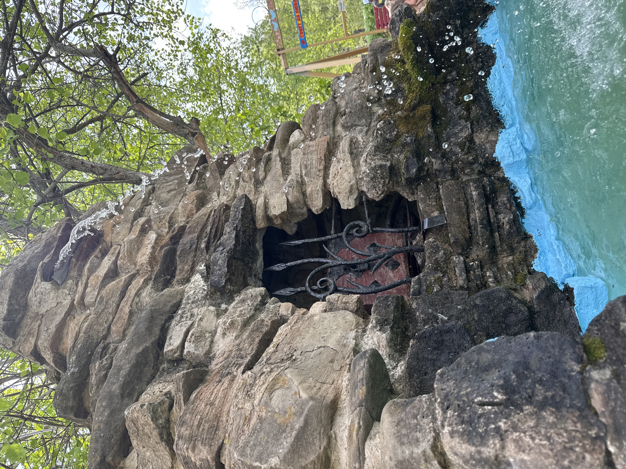 A low-angle, medium shot depicts a stone structure with an opening secured by a wrought iron gate, set against a backdrop of trees and water. The structure appears to be a stone fountain, with water cascading over the top and moss growing on the stone. The water is colored blue.
The opening in the stone structure reveals a red background behind the iron gate. Water droplets are visible along the top of the structure, suggesting water is actively flowing over it.
In the background, above the structure, there's a blurred view of trees and what appears to be a sign or banner with Cyrillic writing, consistent with the location of Chaikovsky, Russia. A section of a red structure, possibly a bench, is also visible.