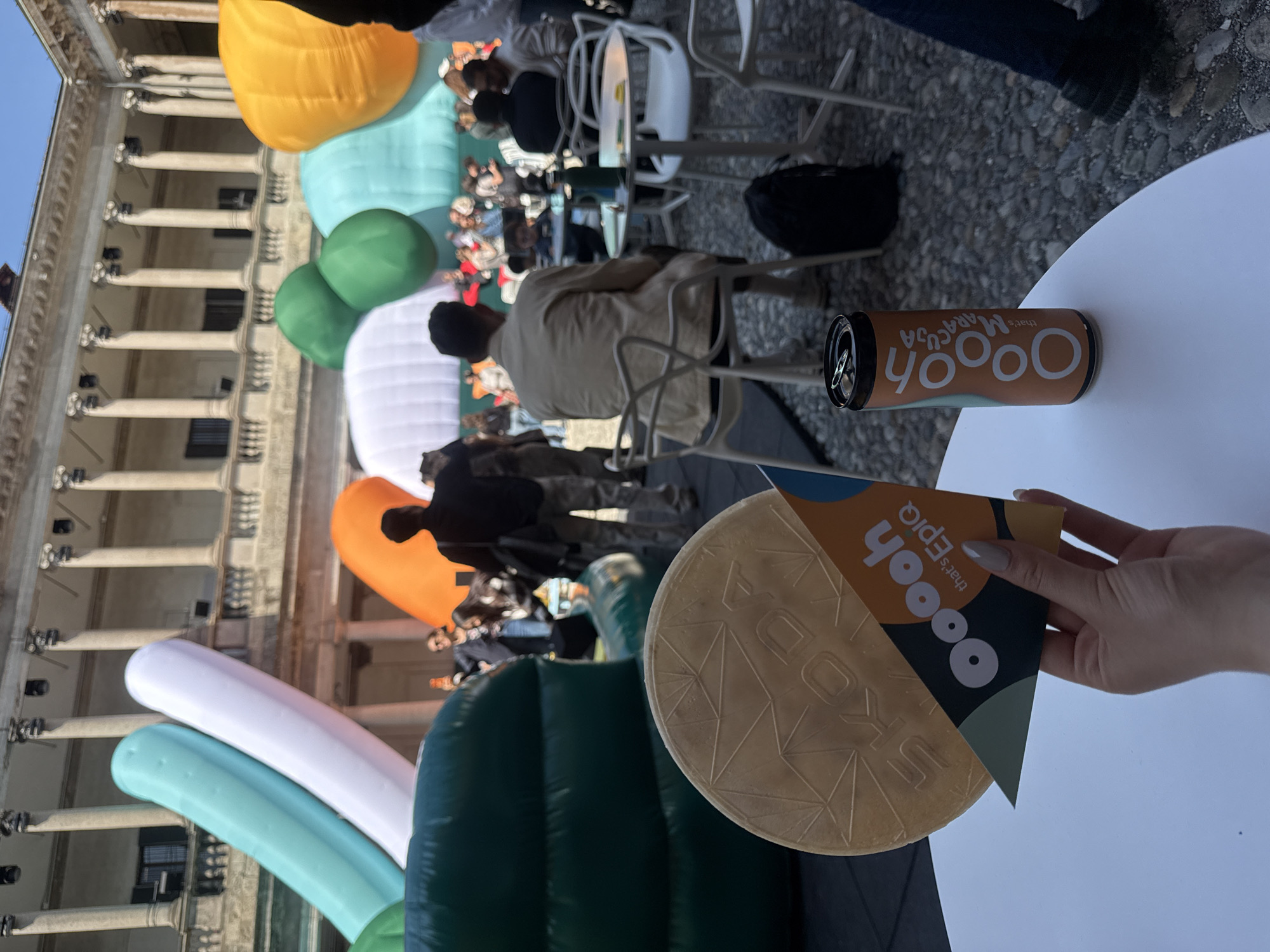 The image depicts an outdoor event taking place in what appears to be a classical European courtyard, likely in Milan, Italy, given the architectural style. The scene is captured from a slightly elevated perspective, looking down onto a white round table in the foreground and the event beyond.
On the white table, a brown beverage can is visible, bearing the text "Oooh that's MARACUJA." A hand, belonging to an unseen person, holds two promotional items: a circular, waffle-like object with embossed lettering, possibly "UKO," and a triangular cardboard piece that reads "Oooh that's Epiq."
The main area of the event features numerous large, colorful inflatable sculptures in organic shapes, rendered in vibrant hues of orange, light blue, green, and white. These modern art installations contrast with the historical backdrop. Several people are scattered throughout the space, sitting at small tables with white chairs or standing, engaged in the event.
In the background, a grand building with prominent columns and arches forms a majestic backdrop, characteristic of a historic palazzo or piazza. The bright sunlight and clear sky indicate it is daytime with pleasant weather. The overall atmosphere appears relaxed and festive, combining cultural architecture with contemporary design elements.