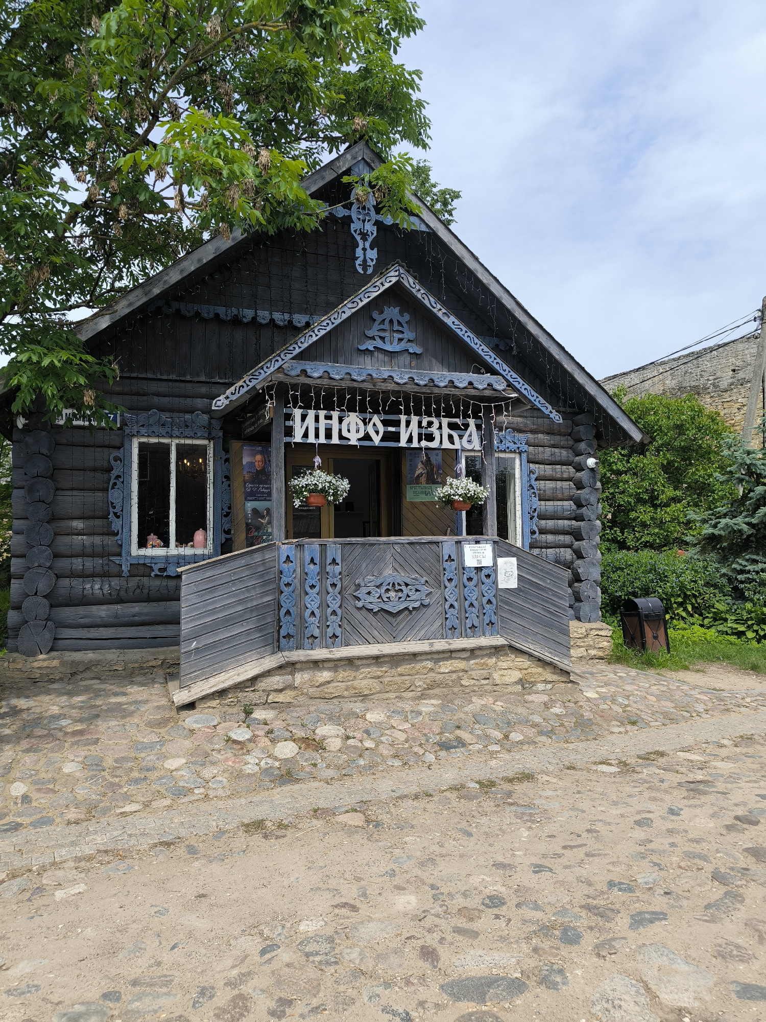 The image shows a traditional Russian wooden house, or izba, in Pechory, Russia. The izba has a dark wood exterior with intricate blue wooden carvings around the windows and eaves. A sign above the entrance reads "ИНФО-ИЗБА" (INFO-IZBA). There are hanging flower baskets and decorative lights along the porch. A cobblestone path leads up to the entrance, and a dirt path is visible in the foreground. Green trees and foliage surround the building, and a stone building is visible in the background. The sky is partly cloudy. No people are clearly visible in the image, though a person's arm can be seen through one of the windows. The scene appears to be a historical or cultural landmark.