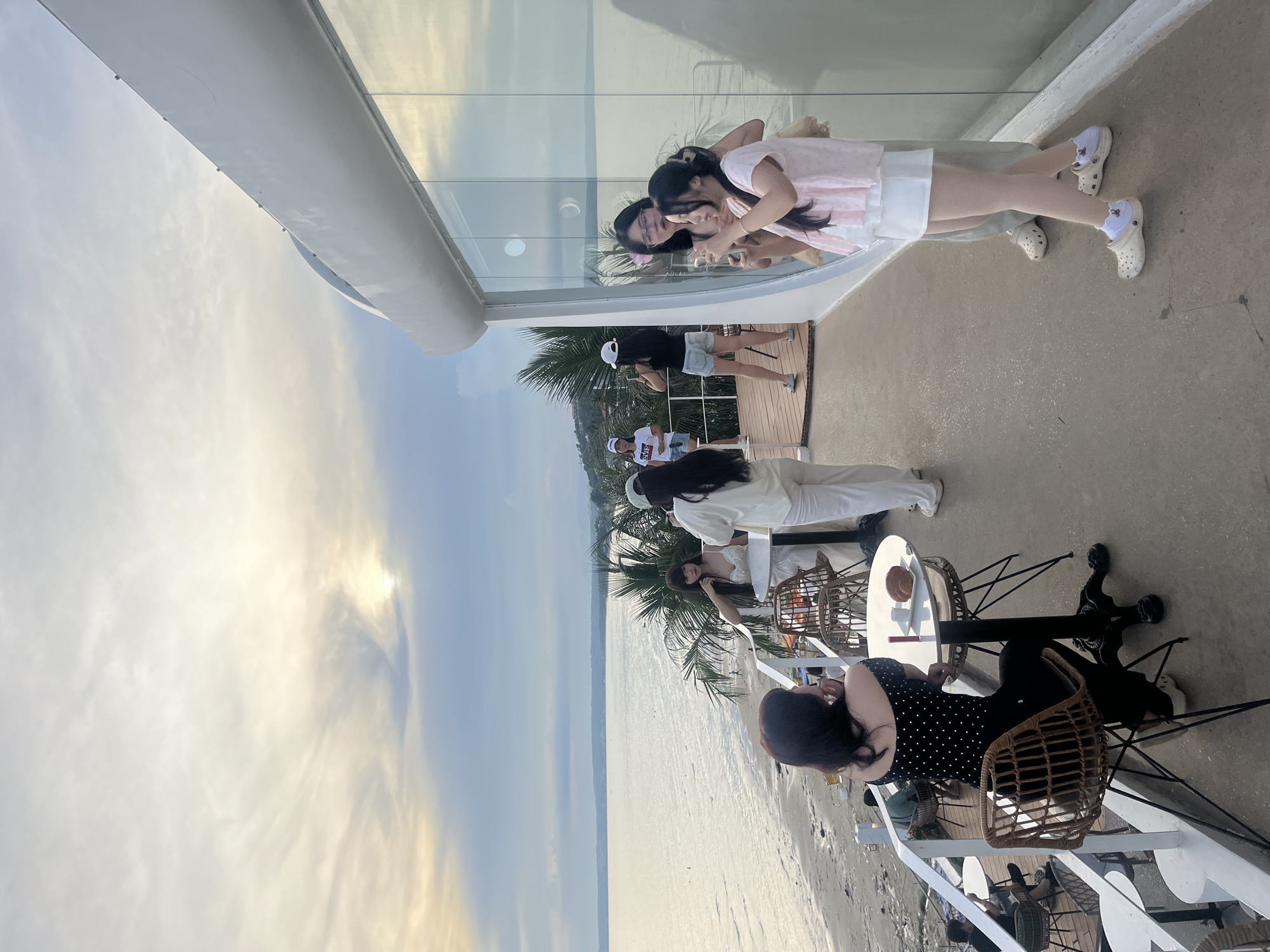 The image depicts a group of people on a deck overlooking a beach and the ocean. The setting appears to be a beachfront cafe or restaurant in Mũi Né, Vietnam, given the tropical foliage, sandy beach, and the style of the architecture.
In the foreground, a woman with dark, curly hair is seated at a round table, facing away from the camera. She is wearing a black off-the-shoulder top with white polka dots and black leggings. On the table in front of her are a red object, possibly lipstick, and a pastry. A wicker chair is pulled up to the table.
Further back on the deck, another group of people are interacting. Two women, one wearing glasses and a light-colored top, and another in a pink and white outfit, are taking photos of each other with their phones. Behind them, to the left, a woman in a white outfit is standing near a table, and another woman in a white t-shirt and denim shorts is also visible. A white railing separates the deck from the beach below.
The environment includes a sandy beach with some scattered debris and a calm ocean. Palm trees and other tropical vegetation are visible along the coastline. The sky is partly cloudy with the sun partially obscured, suggesting it is either sunrise or sunset. The building facade features a large, curved, white structure with extensive glass windows reflecting the sky and the surrounding scene. The deck itself is concrete.