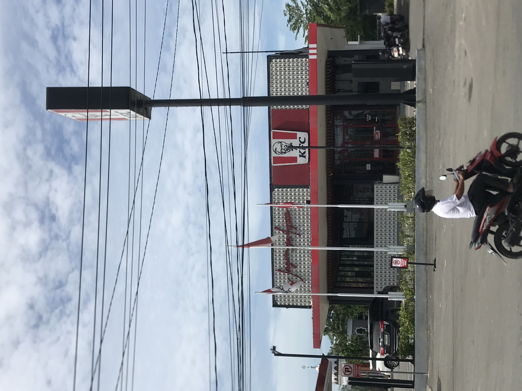 The image depicts an outdoor scene in Lhoksukon, Indonesia, featuring a KFC fast-food restaurant under a partly cloudy sky during daylight hours. The KFC building is characterized by its red and white facade, accented with a grid-patterned upper section. A tall signpost with the iconic red and white KFC logo, including Colonel Sanders and the "KFC" text, stands prominently near the building.
Numerous black power lines stretch across the upper portion of the frame. In the foreground on the right, a person wearing a white shirt and a black helmet is seated on a red and black scooter or motorcycle, facing away from the viewer. The ground is paved, resembling a road or parking lot.
Several small red and white flags are positioned in front of the restaurant. One flag clearly displays Arabic script which reads "لا إله إلا الله" (La ilaha illallah - There is no god but Allah). Another flag appears to have Latin script that is not entirely legible. A "No Parking" sign, denoted by a circled 'P' with a diagonal line through it, is visible on the paved area. In the background, a black SUV and another motorcycle are parked or moving.