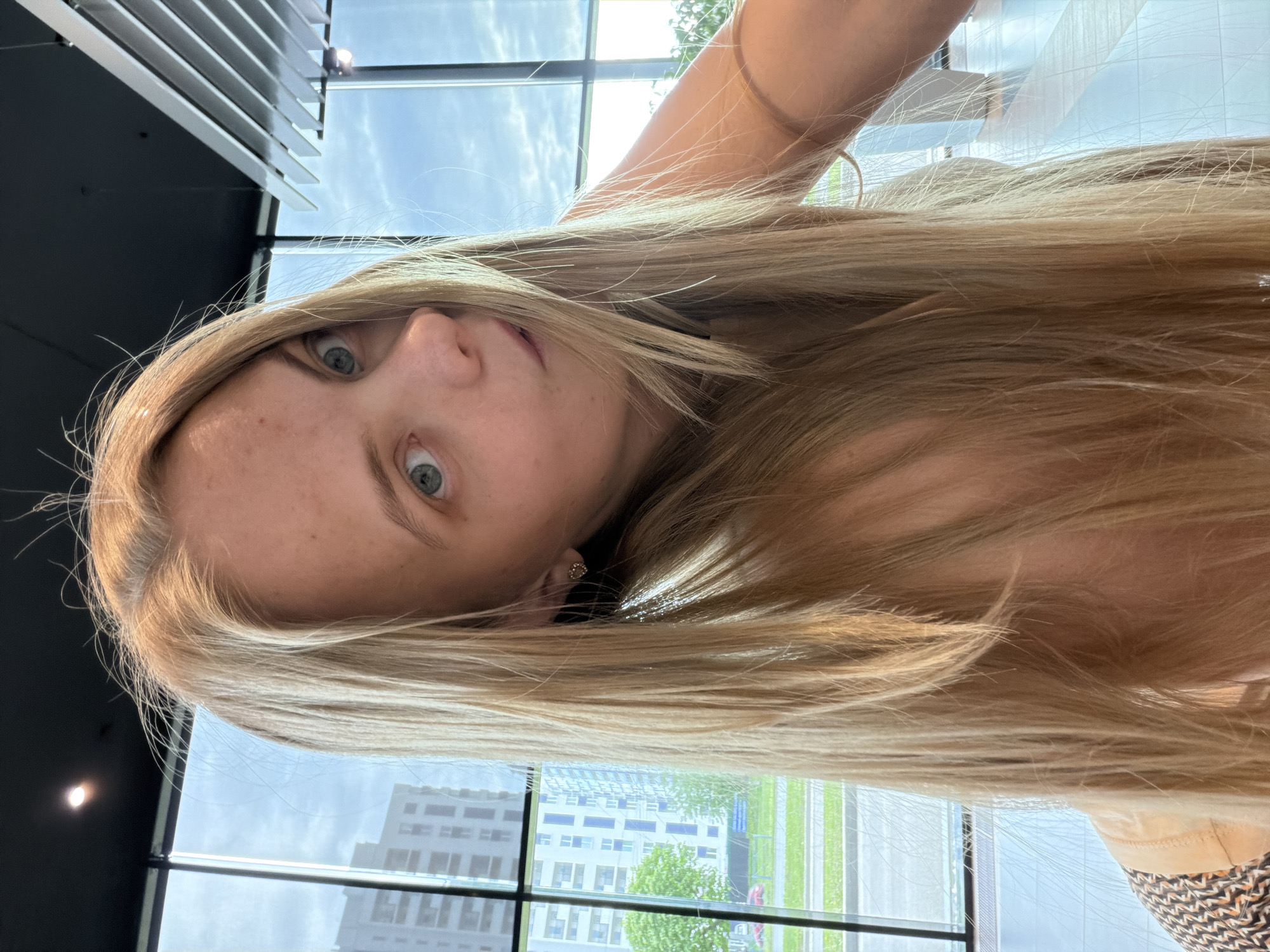 A medium-shot, slightly angled portrait of a light-skinned woman with blonde hair, taken indoors, with a window in the background revealing an urban environment. The woman's face is centrally positioned, showing light blue eyes and fair skin. Her blonde hair is disheveled and covers parts of her body. The background shows a building exterior and glimpses of green vegetation. Given the location of Izhevsk, Russia, visible through the window, the environment appears to be an office interior. The setting is a modern office building. The woman is looking at the camera.