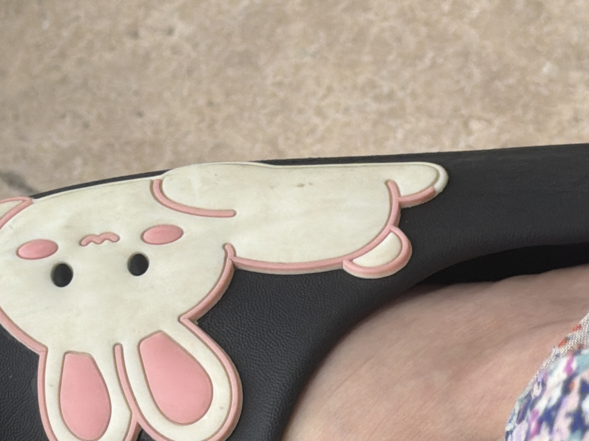This media file shows a close-up view of a black object with a pink and white bunny charm attached. The bunny is depicted with large eyes, a small wavy mouth, and pink inner ears and feet. The black object appears to be made of a textured material, possibly leather, and has a curved shape. A person's forearm, with visible skin texture and veins, is resting on the black object, partially covered by a colorful, patterned fabric, possibly clothing. The background is a blurred, light-colored textured surface, resembling concrete or stone. The lighting suggests it is daytime, with natural light illuminating the scene. There is no visible text in the image. The overall impression is a casual, everyday moment, possibly the charm attached to a bag, shoe, or accessory. The location context is Tbilisi, Georgia.