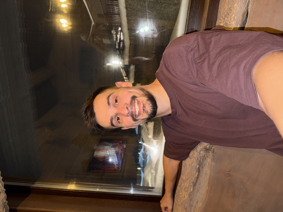 An adult male with short dark hair, a beard, and a mustache is depicted in a selfie, smiling directly at the camera. He is wearing a dark reddish-brown short-sleeved t-shirt with the embroidered text "TWØ" on the left chest. His left arm rests on a textured, light brown wooden or stone-like counter below a large window framed in dark wood. Through the window, a nighttime winter scene is visible, featuring snow-covered grounds and chalet-style buildings with pitched roofs and balconies, illuminated by artificial lights. A distinct reflection of a burning fireplace is visible in the upper left portion of the windowpane. The scene takes place within a lodge or restaurant, likely located at the Bobrovy Log Funpark and Ski Resort in Krasnoyarsk, Russia.