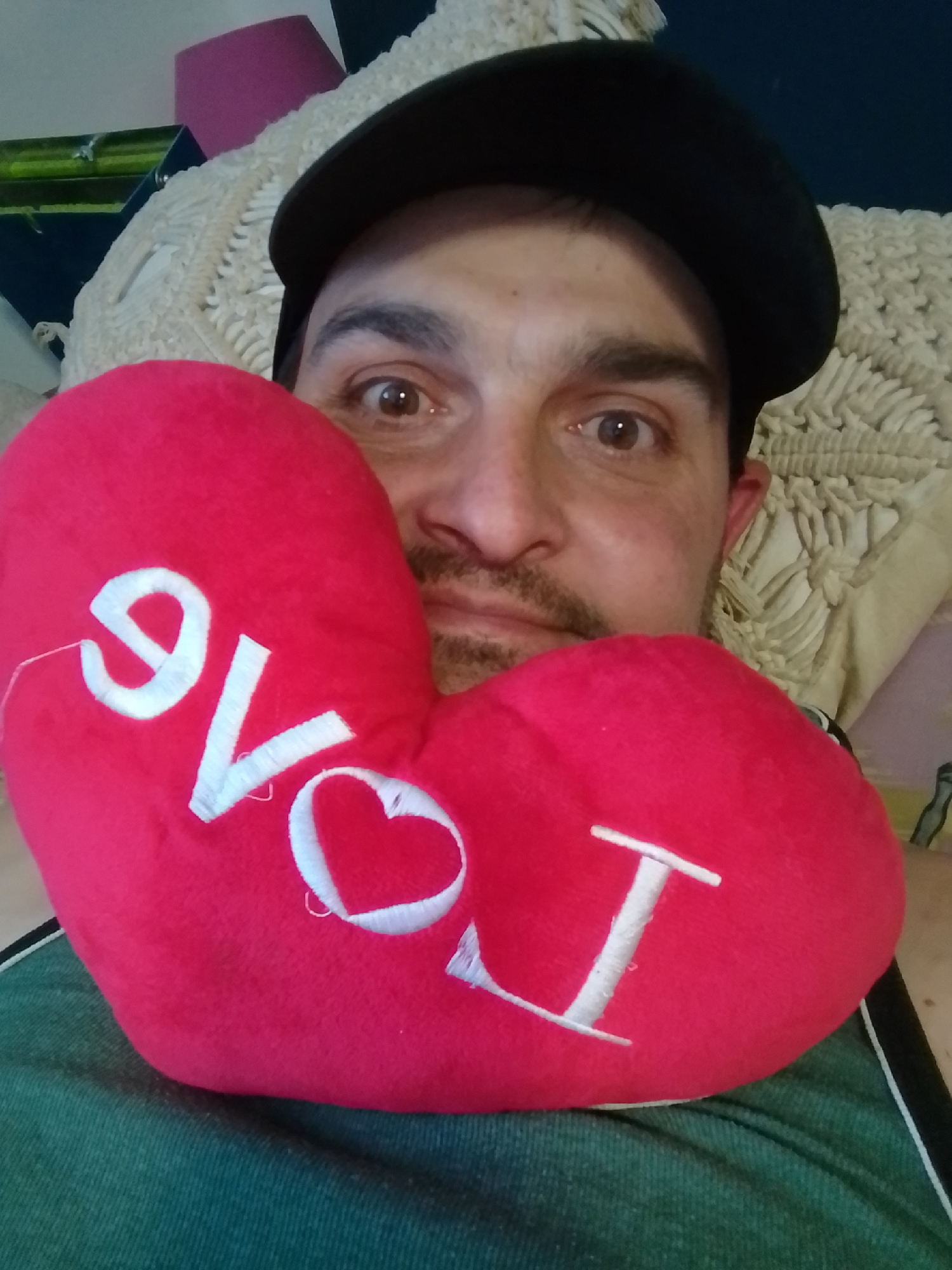 A person wearing a black cap is holding a red, heart-shaped stuffed toy with the word "LOVE" embroidered in white. The person's face is partially obscured by the toy, with their eyes and nose visible. They have brown eyes and a goatee. The background consists of a cream-colored textured surface and a dark blue surface, possibly a wall or furniture. There is also a pink object visible in the upper left corner. The scene appears to be indoors, likely in a domestic setting. There is no information available about the location in the city of Wedel, Germany.