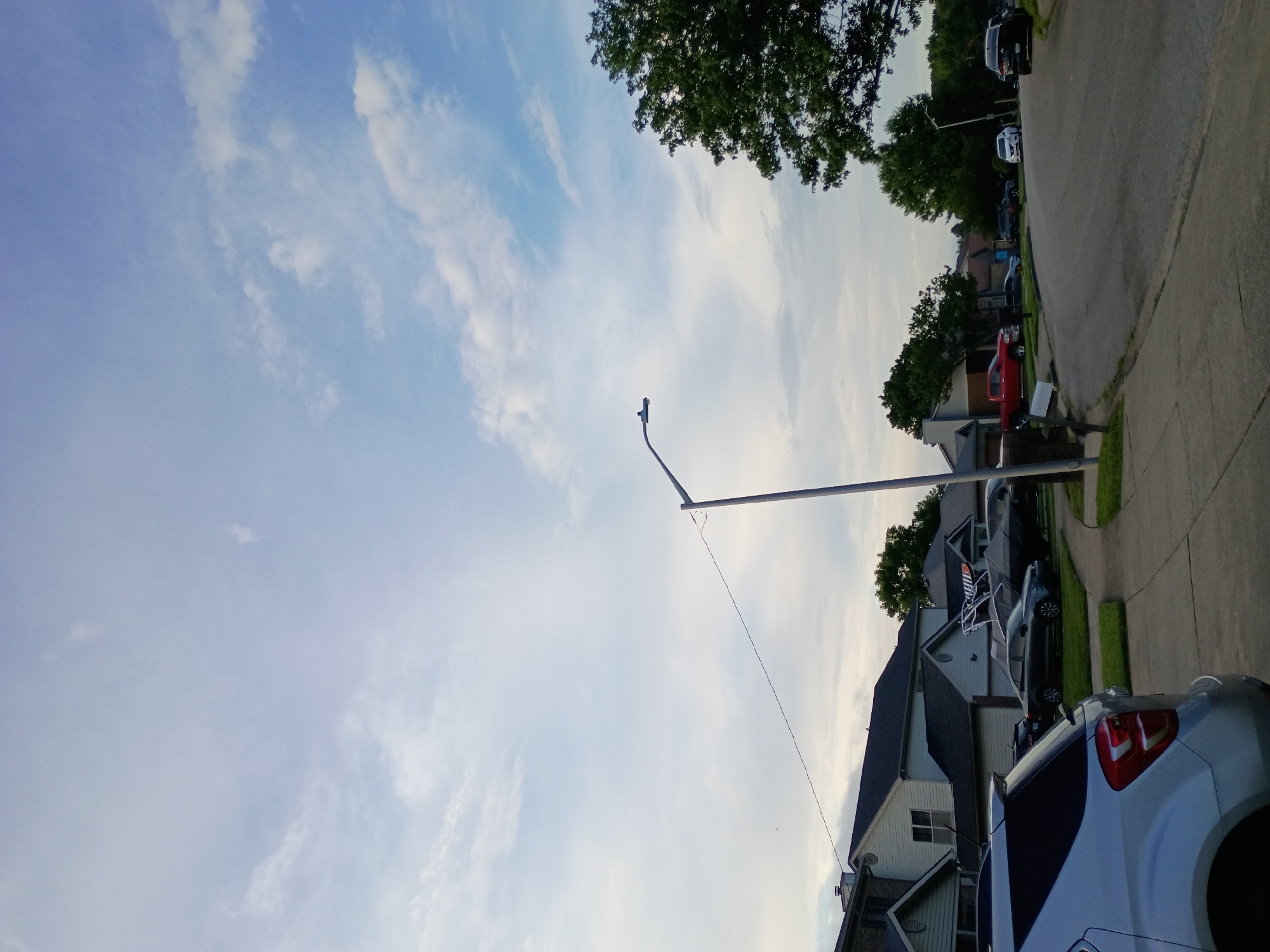The image depicts a suburban street scene in Nicholasville, Kentucky, USA, under a partly cloudy sky. In the foreground, the rear of a silver SUV is visible, parked on a concrete driveway. Beyond the SUV, a sidewalk lines the street. Several houses with pitched roofs and neutral-colored siding are visible along the left side of the street. A red pickup truck and various other vehicles, some covered with tarps, are parked in driveways and along the road. A tall streetlight stands on the right side of the image, its arm extending over the street. Lush green trees and foliage are scattered throughout the neighborhood, with a particularly large tree dominating the right side of the frame. The overall impression is a quiet, residential setting on a day with scattered clouds.