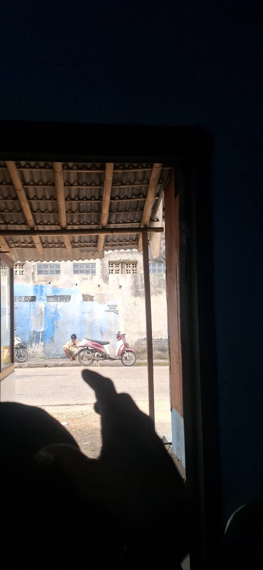 The image depicts an outdoor scene viewed through an opening framed by wooden structures and dark blue walls. In the background, a pale, weathered building with blue paint streaks and patterned ventilation blocks is visible. A person wearing a light-colored shirt and shorts, along with a dark cap, is seated on the ground next to a reddish-pink motorcycle. Another motorcycle is parked to the left of the scene. The ground appears to be a paved surface, possibly a road or sidewalk, with some loose debris. The foreground is dominated by the dark silhouette of a hand casting a shadow on the ground, obscuring details of the immediate surroundings. The lighting suggests it is daytime, with a strong light source casting harsh shadows. The text "DILARANG BUANG SAMPAHI DISINI" (Do not litter here) is faintly visible on the building wall. The location is identified as Cilaku, Indonesia.