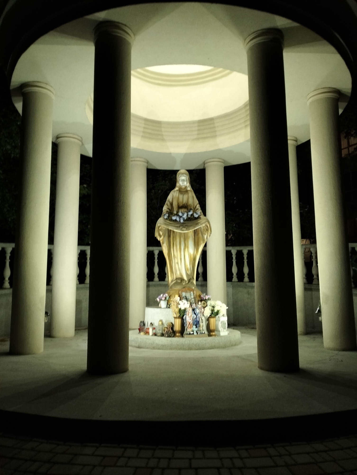 A nighttime shot depicts a statue of the Virgin Mary inside a circular colonnade. The statue, possibly made of gold or a light-colored material, stands on a raised platform adorned with flowers and religious objects. The colonnade consists of multiple columns supporting a round roof with a circular opening at the top. The lighting is subdued, casting shadows from the columns onto the ground. The foreground shows a paved area, and beyond the colonnade, glimpses of foliage suggest a park or garden setting. This is located in Krykhivtsi, Ukraine.