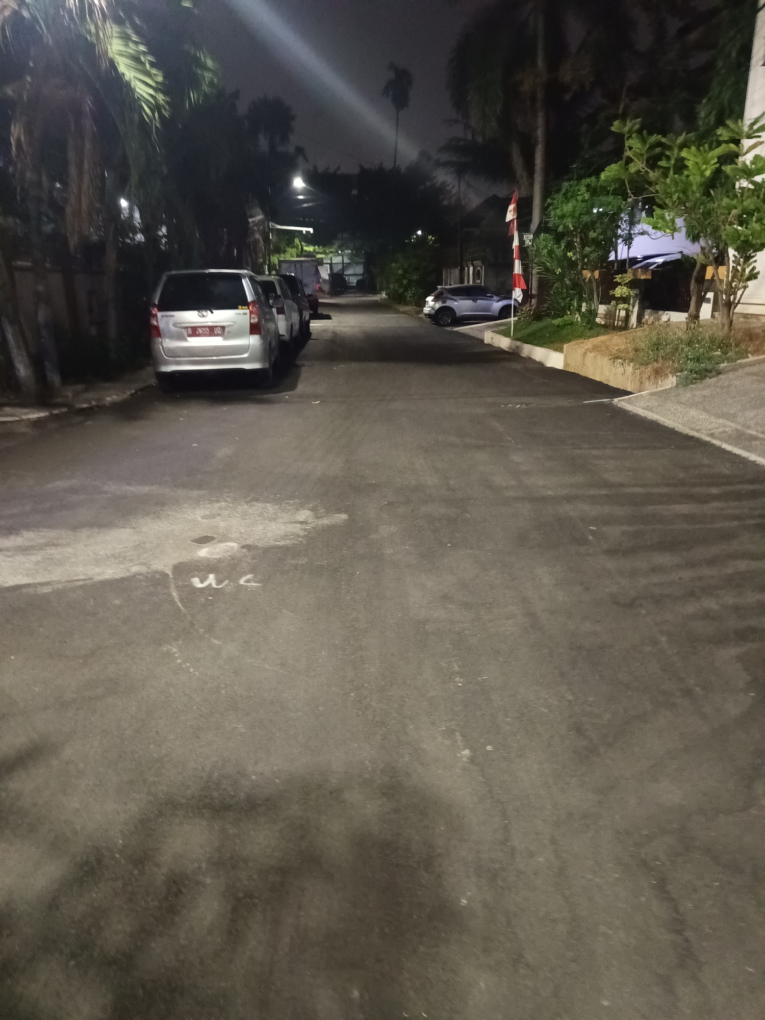 The image depicts a street scene at night in North Jakarta, Indonesia. A silver minivan is parked on the left side of the road, with its license plate visible. Further down the street, another car is parked on the right side. The street is lined with trees and a few buildings are visible in the background. The road surface appears to be asphalt and shows some wear and tear. There are some white markings on the road in the foreground. The overall lighting suggests it is a residential or commercial area with streetlights providing illumination. The atmosphere is dark and quiet, typical of a nighttime scene in an urban environment.