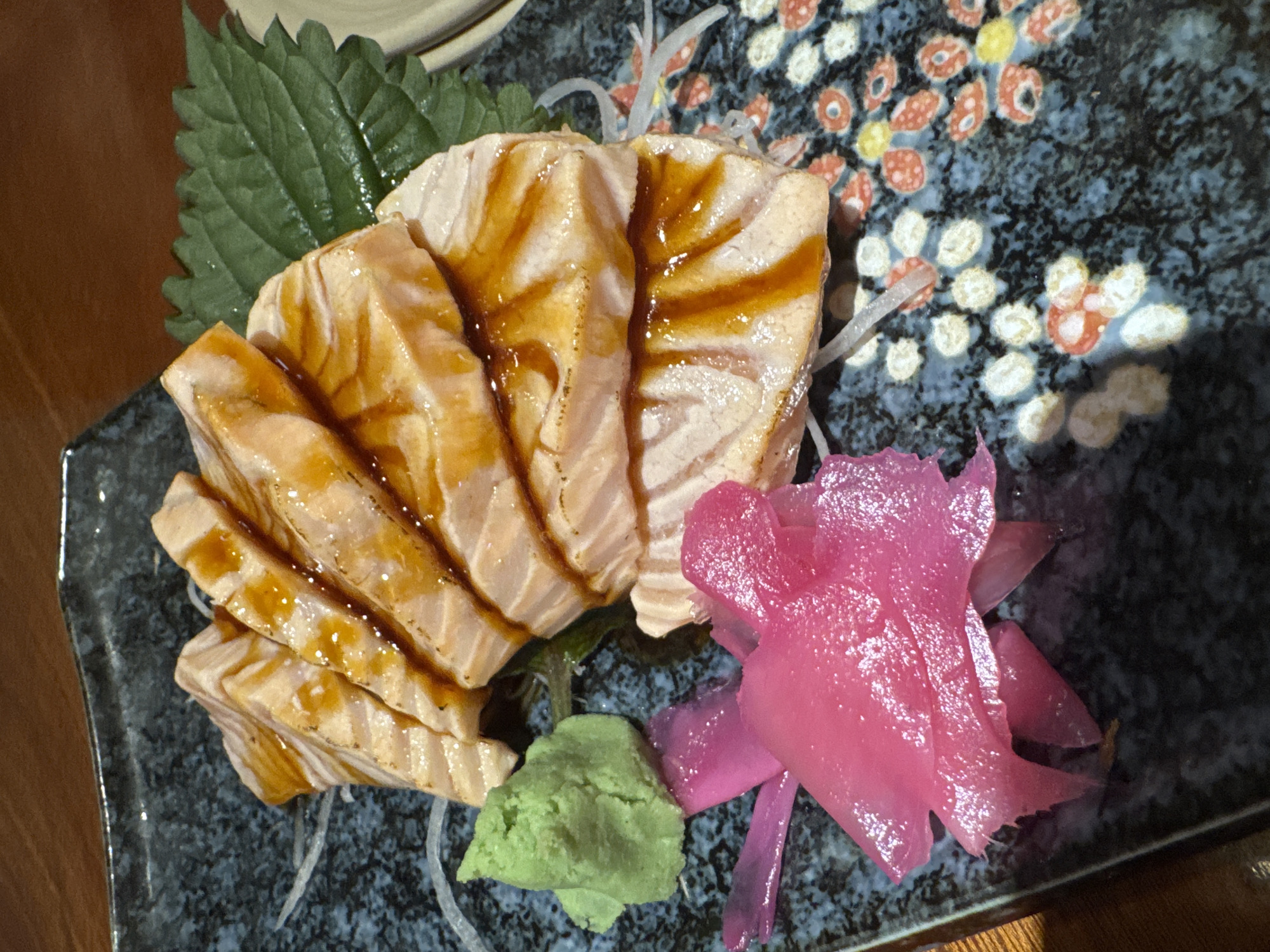 A close-up shot of salmon sashimi on a patterned blue and black plate. The sashimi is arranged with a dark green leaf, a dollop of bright green wasabi, and a pile of pickled ginger. A brown glaze is drizzled across the slices. The plate is placed on a wooden surface. The image does not provide information about Đà Nẵng or Vietnam.