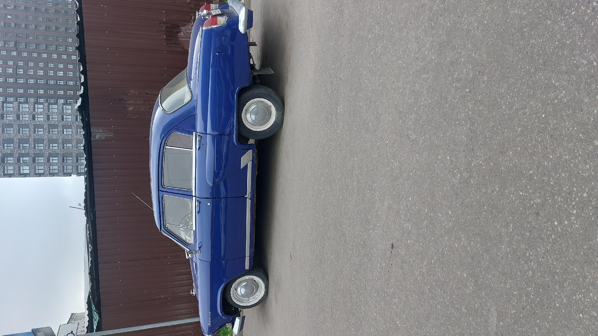The image shows a blue GAZ-21 Volga car parked on an asphalt surface. A tall, modern apartment building stands in the background, along with a brown corrugated metal fence. The scene appears to be in a city environment, possibly Moscow, Russia. No people are visible in the frame. The car seems well-maintained and is a classic example of Soviet-era automobile design. The environment suggests an urban setting with a mix of old and new infrastructure.