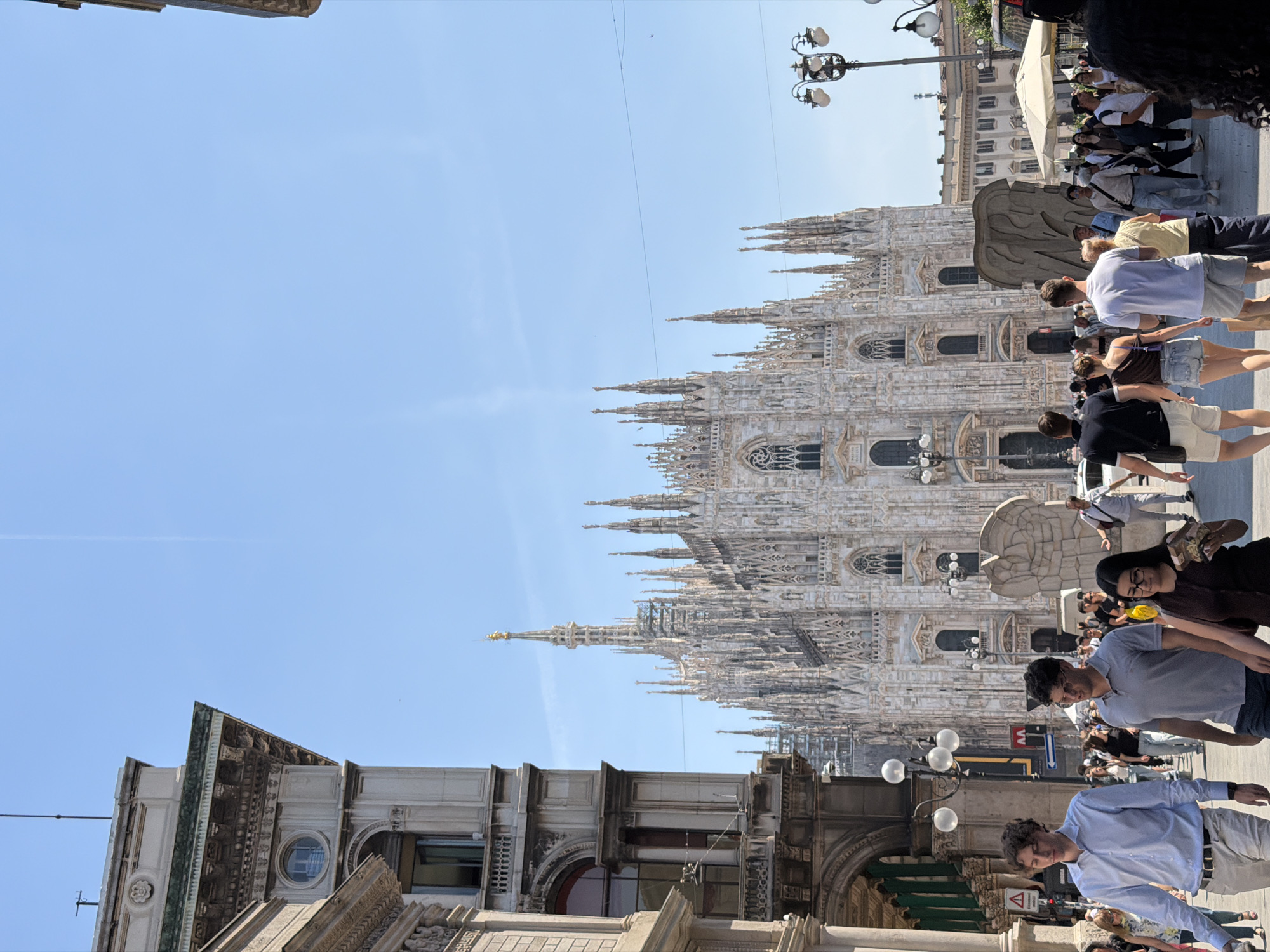 The image shows the Milan Cathedral (Duomo di Milano) in Milan, Italy. The cathedral, a large Gothic-style structure with numerous spires and architectural details, dominates the skyline. In the foreground, people are walking along the street and standing near the building facades. The sky is blue and clear. Street lamps are visible among the buildings and people.