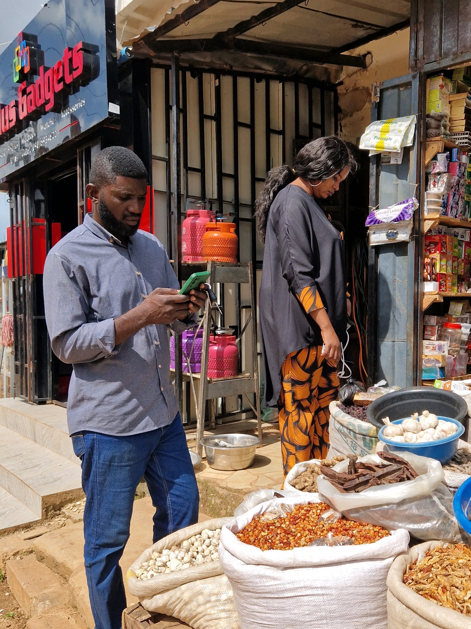 In Jos, Nigeria, a man in a blue collared shirt and jeans stands looking at his phone, holding it in his right hand. Beside him, a woman in a black tunic and patterned orange and black trousers stands with her hands clasped in front of her. In the background, a shop displays various items, including propane tanks. In the foreground, sacks filled with spices and vegetables are visible, along with a blue bowl containing garlic.