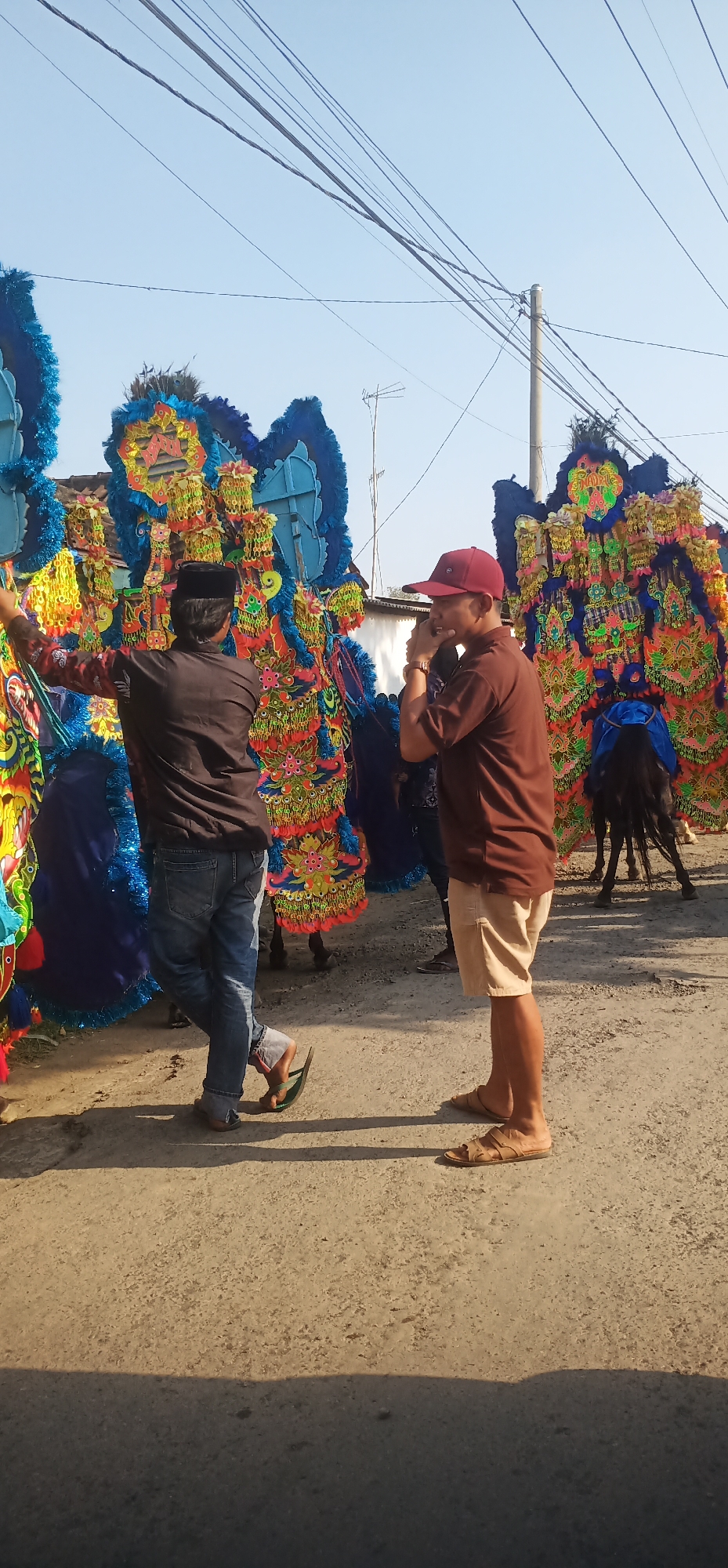 In Kalisalam, Indonesia, two men are standing near a procession of colorful, elaborate costumes. The man on the left, wearing a black cap and brown shirt, faces away from the camera, his left arm extended towards the costumes. He is wearing blue jeans and flip-flops. The man on the right, sporting a red baseball cap and a brown t-shirt, faces left, holding his right hand to his mouth as if speaking or taking a picture. He is wearing tan shorts and sandals. Behind them, large, vibrant decorations, possibly part of a cultural performance or parade, are prominent. One of these decorations has bright blue fringe and intricate patterns in yellow, red, and green. A dark-colored horse with a blue saddle blanket is visible between the men and the decorations on the right. The ground is an asphalt road, with shadows indicating a bright, sunny day. Electrical wires and poles are visible in the background, suggesting an urban or semi-urban environment.