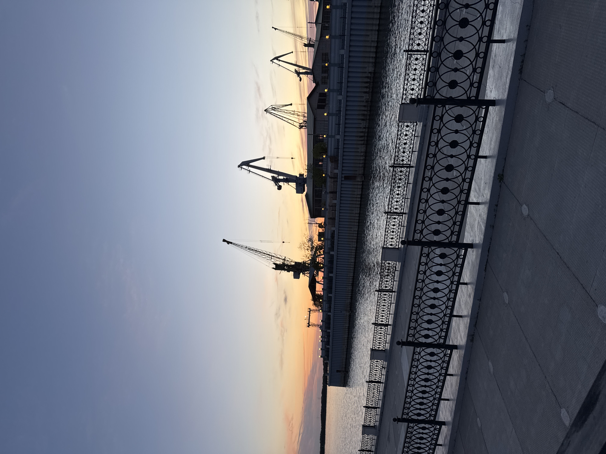 The image depicts a cityscape at dusk in Khabarovsk, Russia. A row of cranes stands silhouetted against a sky gradient of light blue to orange, indicating either sunrise or sunset. These cranes are located near a waterfront area, with a dark, reflective body of water visible between them and the foreground. A decorative black metal railing runs along the edge of a paved walkway, suggesting a promenade or pedestrian area. The walkway extends into the frame, showing its textured surface and occasional circular features. The overall scene conveys a sense of industrial activity juxtaposed with a tranquil urban setting.