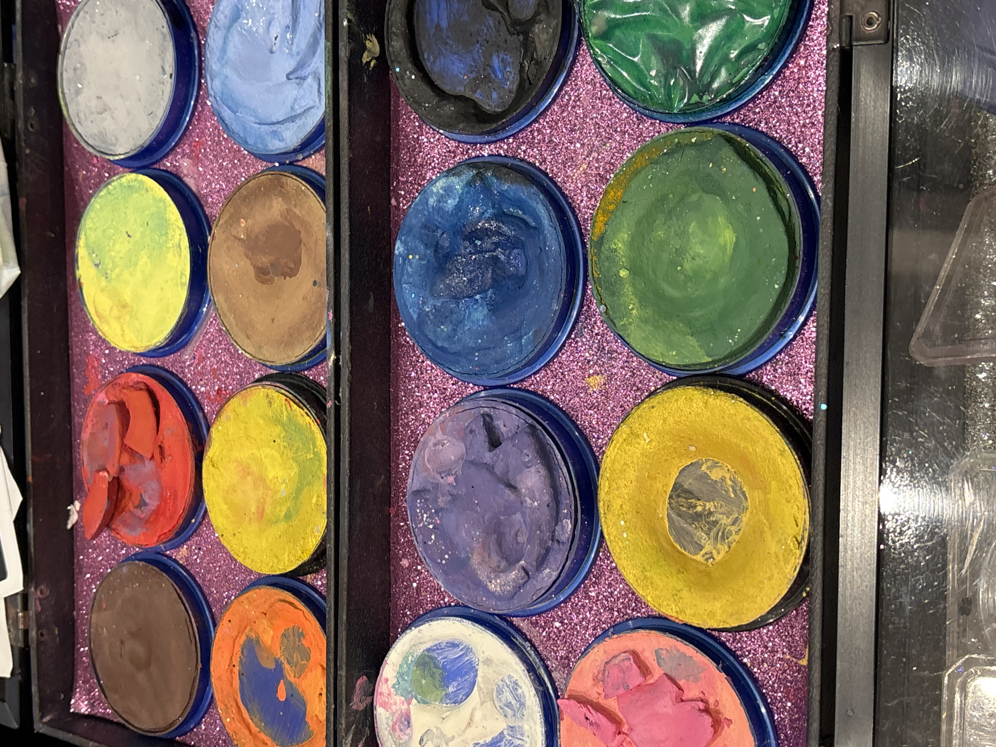 A flat lay overhead shot shows an open makeup palette containing various cream makeup colors. The palette is black with a textured, glittery pink interior. The makeup is arranged in a grid of circular pans.
In the top row, from left to right, are pans of brown, red, yellow, and light gray makeup. The red makeup pan appears partially used, with some of the cream broken into chunks.
The second row, from left to right, contains orange with blue swirls, yellow, brown, and light blue makeup.
The third row, from left to right, shows white with colorful streaks, purple, a darker blue, and black makeup. The purple and blue pans show signs of use, with some glitter visible.
The bottom row, from left to right, includes pink, yellow with a dark center, green, and green with a textured surface. The yellow pan has a distinct circular depression in the center.
The overall condition of the makeup suggests it is frequently used. The image does not provide any information that would suggest it is located in Dubai, United Arab Emirates.