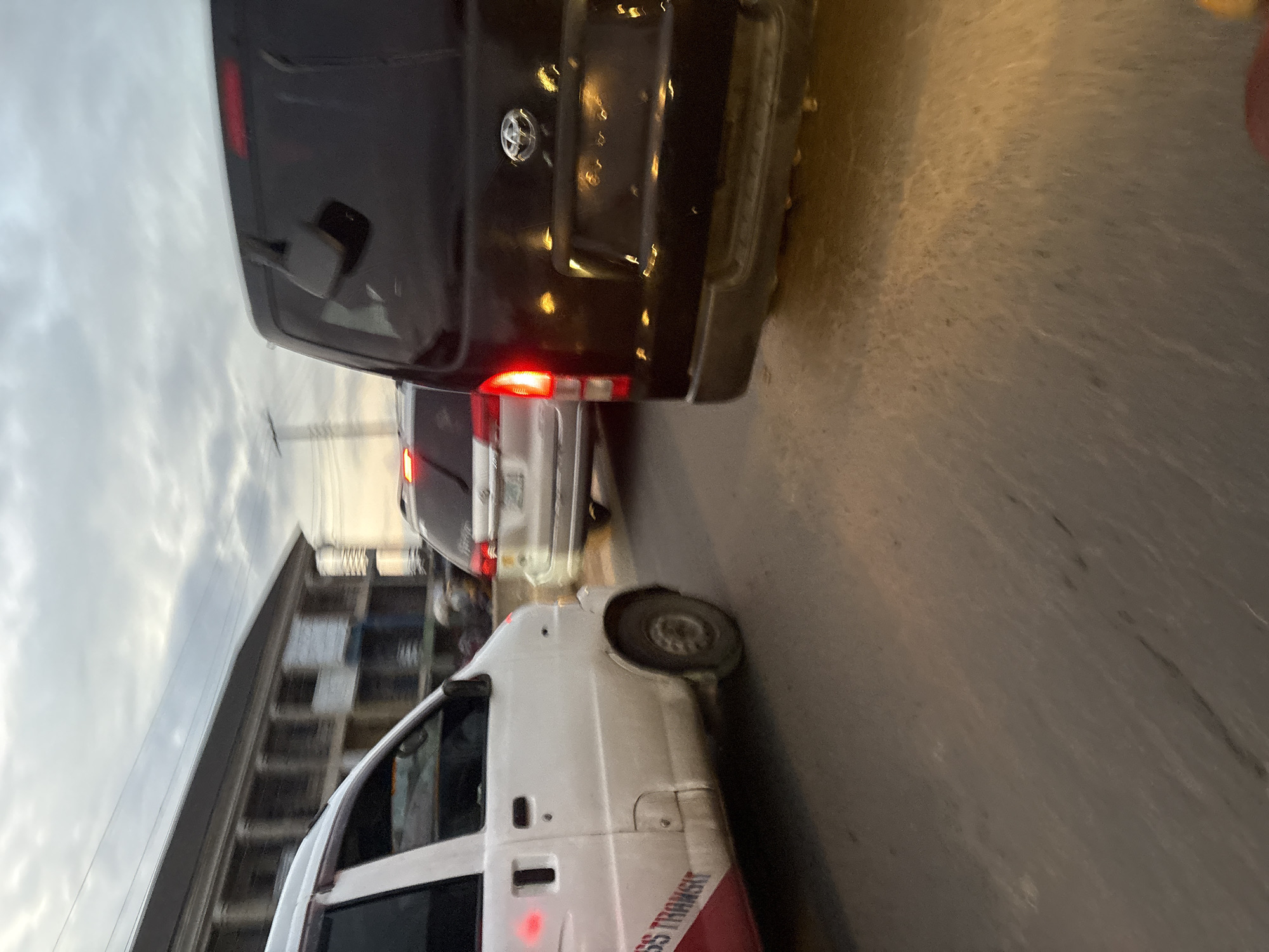 The image shows a street scene in Onitsha, Nigeria, with several vehicles on a road under a cloudy sky. In the foreground, a white mini-van with red and blue lettering that reads "SS TRANSIT" is on the left. To the right of the white van is a dark-colored SUV, possibly a Toyota, with its brake lights illuminated. In the background, another silver SUV is visible. The vehicles appear to be in traffic. Buildings with classical architectural elements, including columns, are visible behind the vehicles on the left side of the image. The road surface is asphalt. The overall lighting suggests it is either early morning or late afternoon.