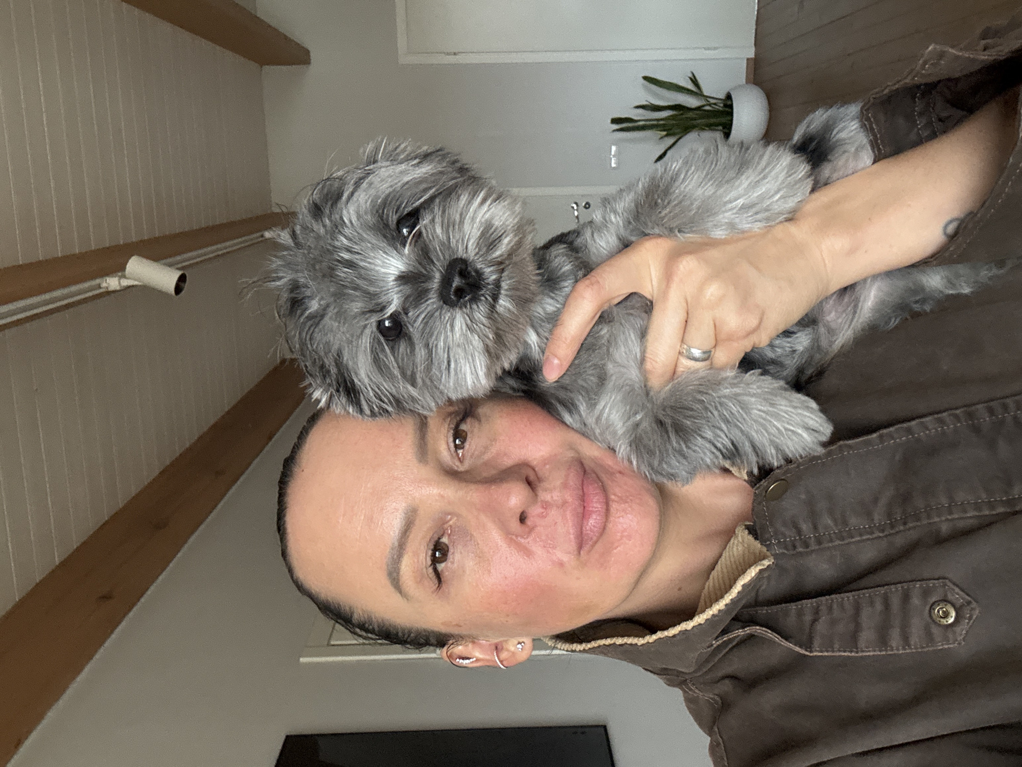 A woman is holding a small, grey dog in her arms. The woman has dark hair pulled back, fair skin, and is wearing a brown jacket. She is looking directly at the camera. The dog is also looking at the camera with its head resting on the woman's shoulder. The background shows the interior of a room with a sloped ceiling and wooden beams. There is also a plant in a white pot visible.
There is no indication of Chaikovsky, Russia.