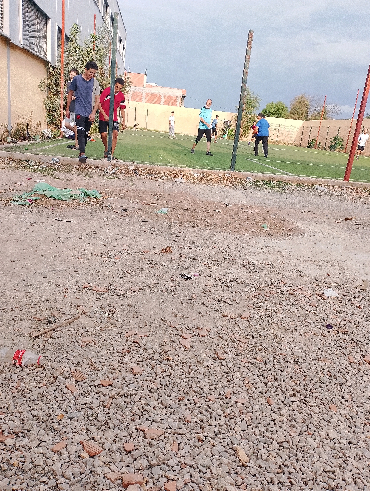 The image depicts an outdoor scene on a cloudy day, likely in Bordj Menaiel, Algeria, with a focus on a dilapidated outdoor sports field. In the foreground, the ground is covered with loose gravel, scattered pieces of broken brick and tile, and some discarded green plastic sheeting. A single plastic water bottle with a red label lies on its side amongst the gravel.
Further into the image, an artificial turf sports field is visible, marked with white lines. Several individuals are present on the field, seemingly engaged in some form of athletic activity or leisure. Two young men in the mid-ground are walking towards the viewer, one in a blue and gray t-shirt and black pants, the other in a red t-shirt and dark shorts, both appearing to be smiling and in conversation. Other individuals, dressed in athletic wear, are scattered across the field, including a person in a light blue shirt and black shorts, and another in a blue t-shirt and black pants. A person in a white t-shirt and dark shorts is visible on the far right of the field.
The background features a building with a light-colored facade and windows, adjacent to a chain-link fence. Red-orange brick buildings are also visible in the distance. The overall environment suggests an urban setting, possibly a public space undergoing some level of disrepair or construction, with a sports facility integrated into it. The sky is overcast, indicating a potentially cool or humid day.