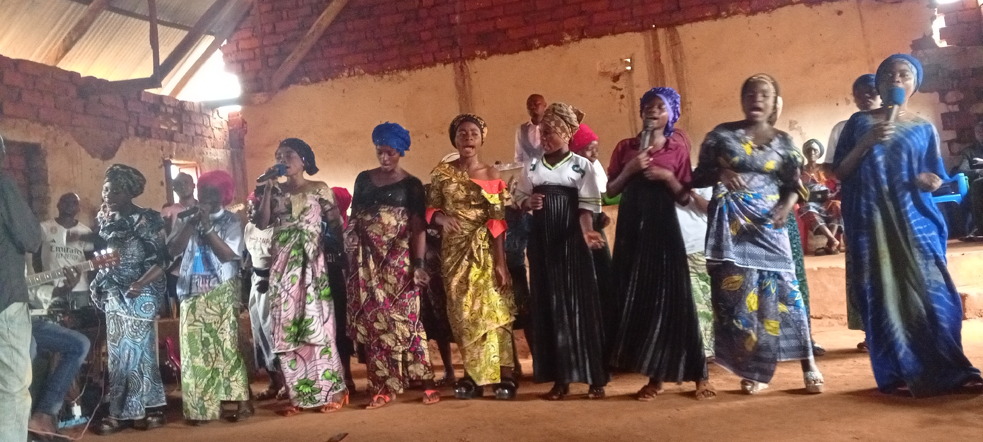 In Ngalula, Democratic Republic of the Congo, a group of approximately 14 people, mostly women, are singing in what appears to be a church or community building with exposed brick walls and a corrugated metal roof. Several of the women are holding microphones and leading the singing. The women are wearing a variety of colorful traditional African clothing. A man with a guitar stands to the side providing musical accompaniment. Some people are seated and watching. The ground is dirt or packed earth.
