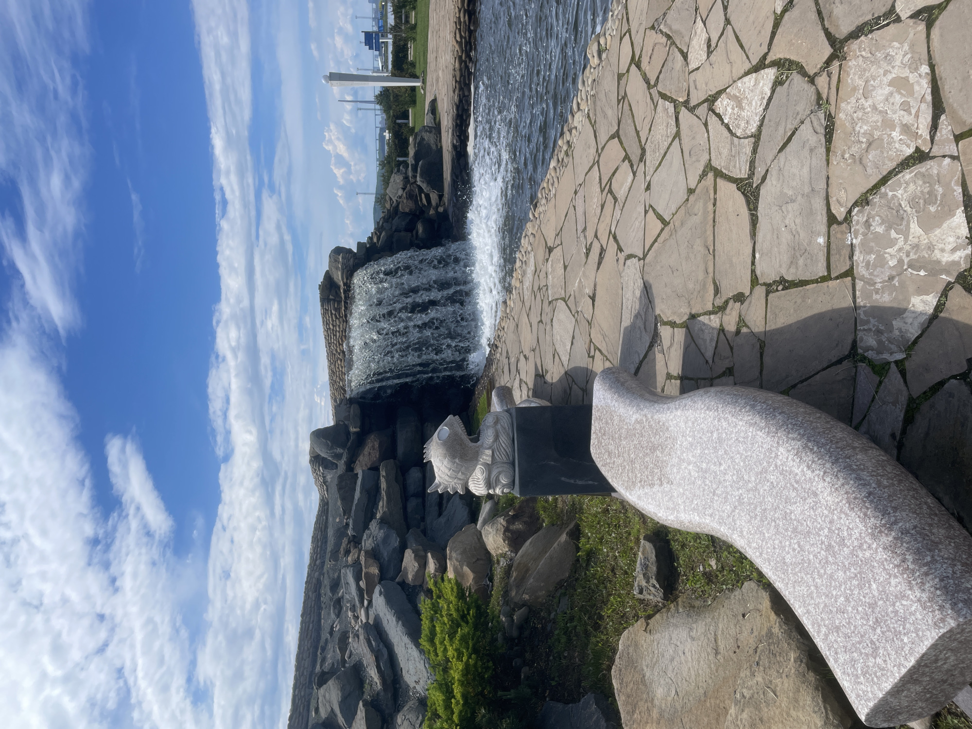 In Russky, Russia, a picturesque scene unfolds with a cascading waterfall as the central element. The waterfall, a man-made structure constructed with rocks, tumbles into a body of water. To the left of the waterfall, a stone sculpture of a fish, with an open mouth and stylized fins, sits on a dark rectangular pedestal. In the foreground, a curved, light-colored granite bench is positioned on a pathway paved with irregular flagstones. Beyond the waterfall and water, a neatly manicured lawn stretches towards distant structures and a bright blue sky dotted with white clouds. The overall ambiance is serene and inviting, suggesting a public park or recreational area.