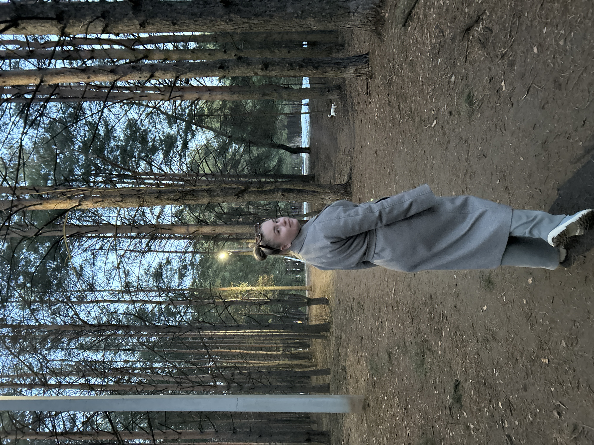 A woman stands on a dirt path in a forest in Chaikovsky, Russia. She wears a gray coat and gray pants with light-colored shoes. The forest consists of tall, thin trees. In the background is a small, white dog. A streetlight is visible in the background.