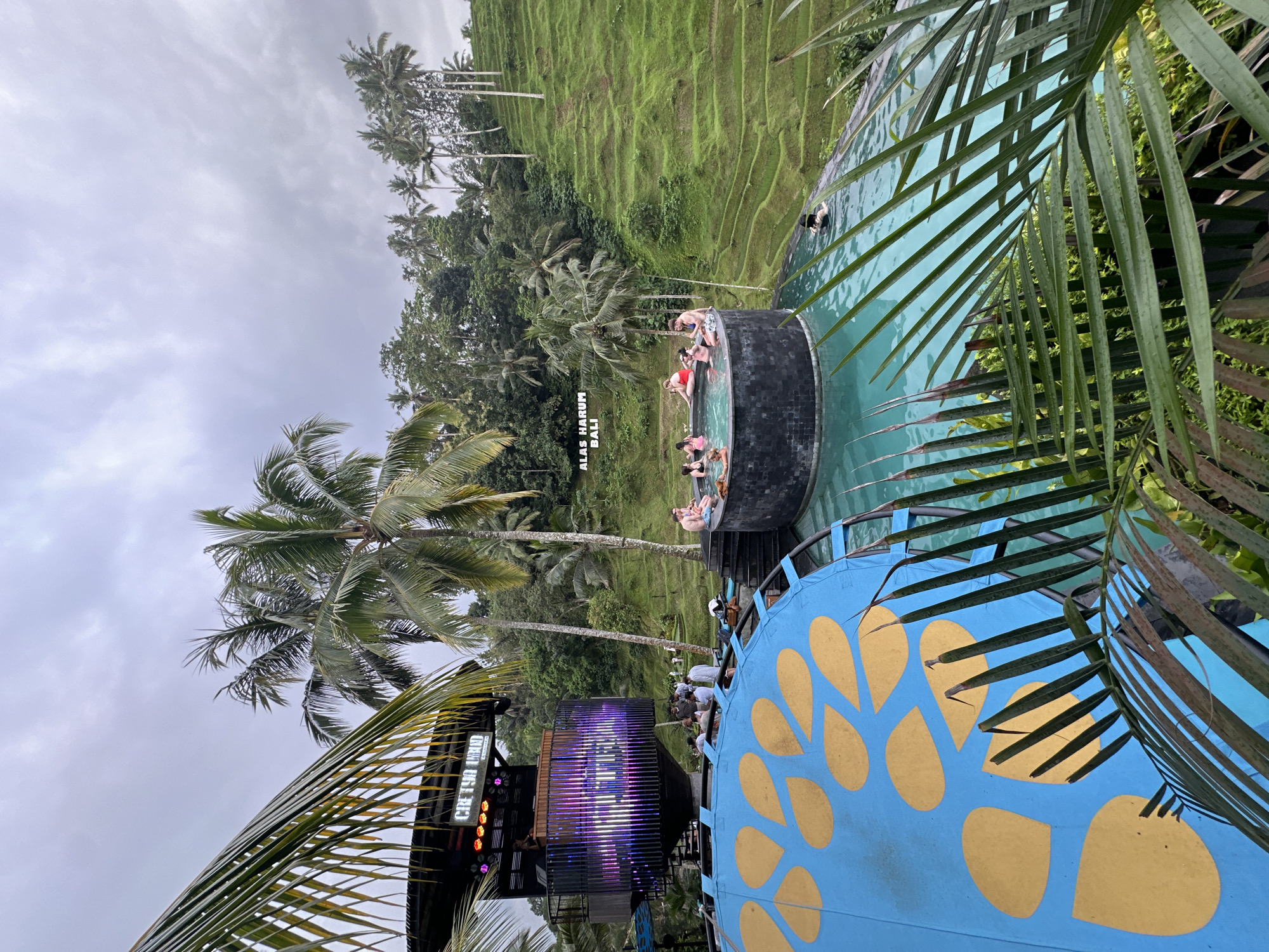 This photo, taken in Tegallalang, Indonesia, captures an outdoor scene featuring a multi-tiered swimming pool surrounded by lush tropical vegetation. The pools are occupied by people, some lounging and others swimming. Nearby, a stage or structure with purple lighting is visible. A blue and gold structure with decorative patterns is located in the foreground. The environment consists of palm trees and terraced rice paddies, characteristic of the Tegallalang area. The sky is overcast.