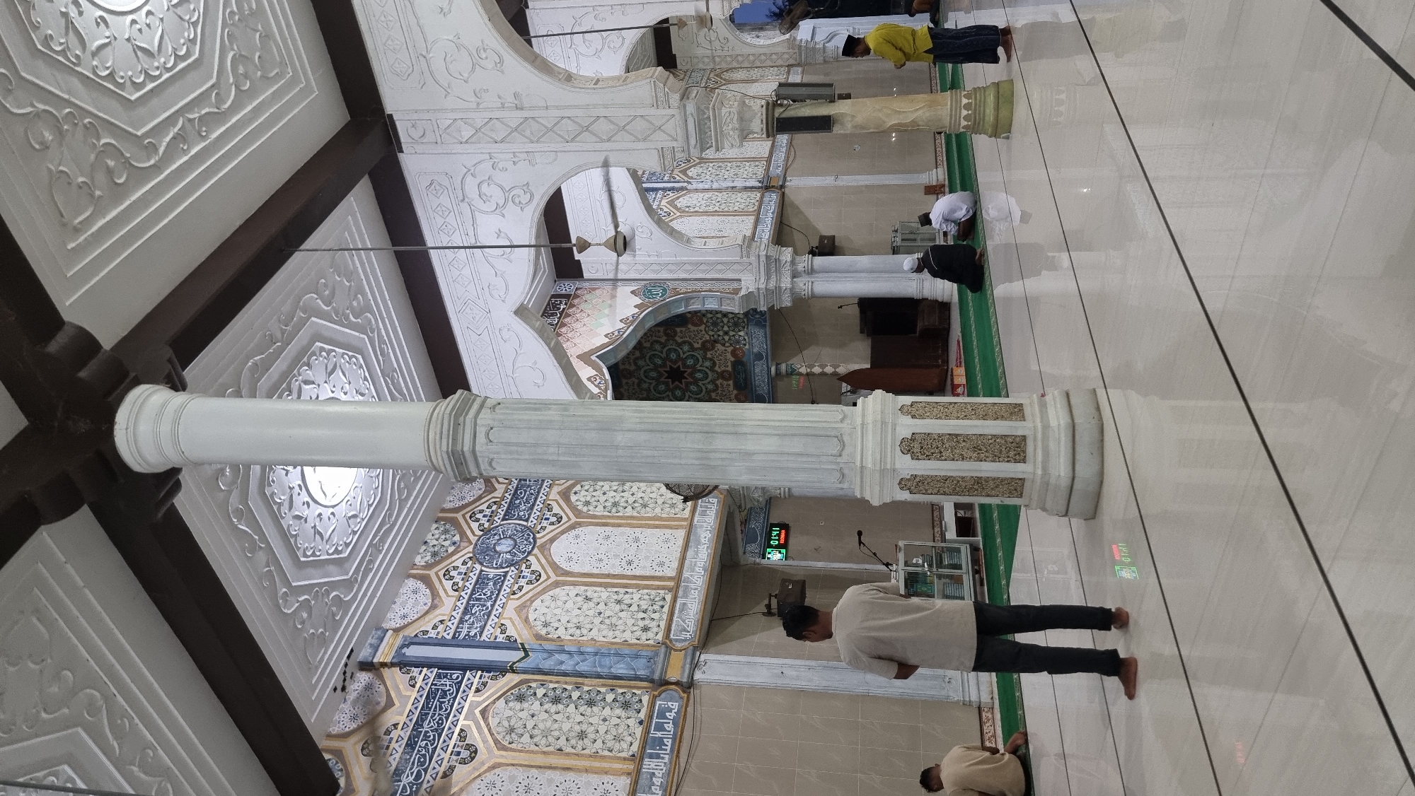 A wide-angle shot captures the interior of a mosque in Meunasahlhok, Indonesia, with a focus on the ornate architecture. In the foreground, a large, fluted white pillar with decorative gold accents stands prominently. The floor is made of polished white tile with thin black lines marking prayer rows, and the reflections of the ceiling and pillars are visible.
To the left, a man wearing a light-colored t-shirt and dark pants stands with his back to the camera, facing the front of the mosque. Further left, two men are seated on green prayer mats, facing the same direction.
The walls are adorned with intricate blue and gold patterned tiles, featuring geometric designs and Arabic calligraphy. A digital clock on the wall displays "01:41." Behind the seated worshippers, a tall glass cabinet with religious books is visible.
Towards the center background, a large archway reveals the Mihrab, the prayer niche, decorated with a starburst pattern and Arabic calligraphy. A ceiling fan is visible in the upper right portion of the frame.
To the right, two men are kneeling on prayer mats. One wears a dark thobe and a white skullcap, and the other wears a white thobe and skullcap. Further to the right, a man in a yellow t-shirt and dark pants stands observing. The ceiling is white with elaborate decorative molding and dark wooden beams. The overall atmosphere suggests a quiet moment of prayer or reflection within the mosque.
