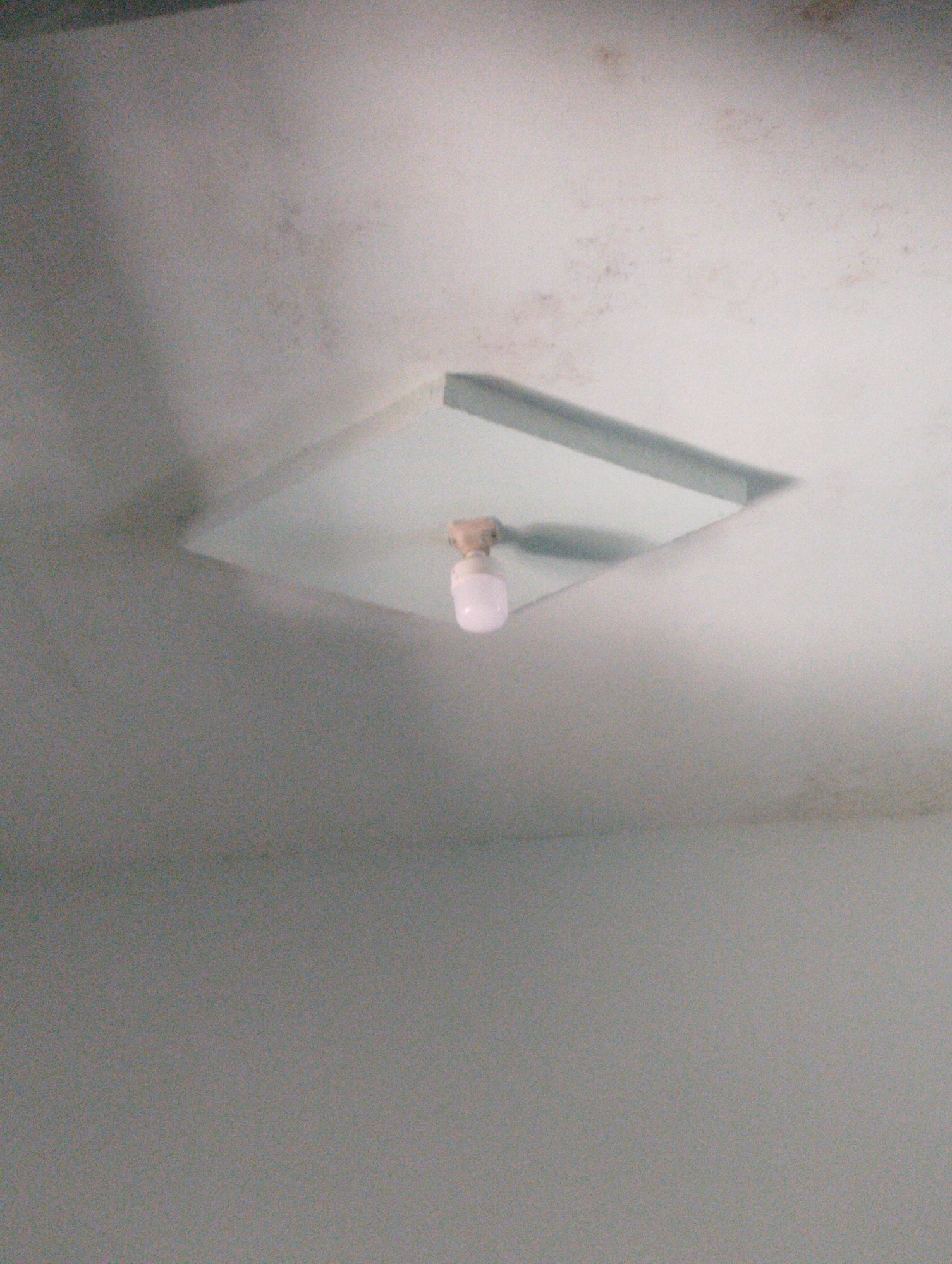 The image shows a ceiling light fixture with a single, pink-tinted light bulb. The fixture is square and appears to be made of metal and glass. The ceiling itself is a light color, possibly white or off-white, and shows some signs of staining or discoloration, particularly in the upper portion of the image. The lighting in the room is dim, with shadows cast by the light fixture and the ceiling itself. The image is taken from a low angle, looking upwards. There are no people or any other objects visible in the image. The location in Wanasaba Daya, Indonesia, is not discernible from the visual information present in the image.