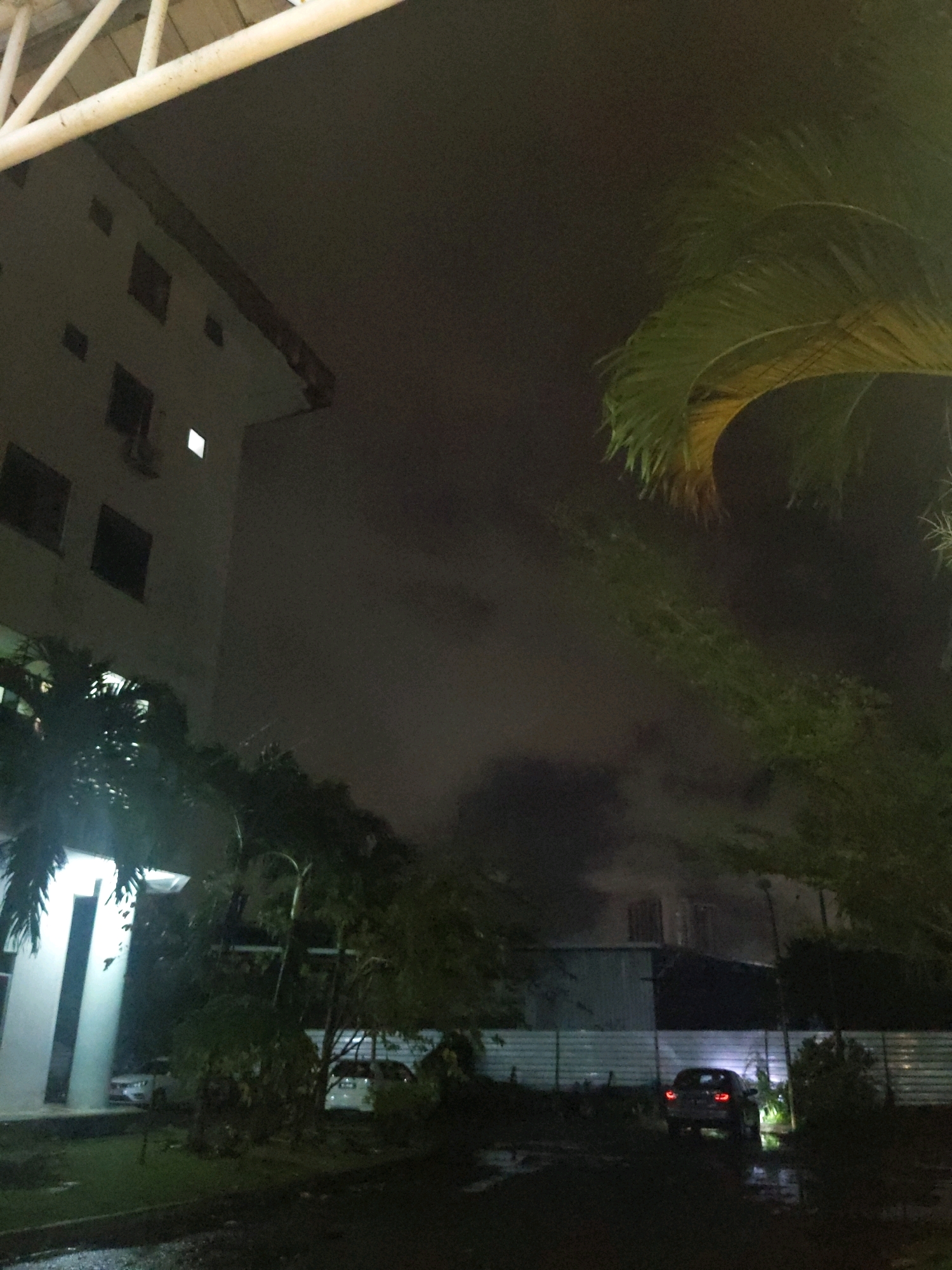 An exterior, low-light shot captures a modern building in Kolombong, Malaysia, at night. The building's façade is illuminated by a few scattered lights, with some windows appearing dark and others faintly lit. A prominent white overhang supported by beams stretches across the upper portion of the frame. Lush green palm fronds frame the right side of the image, their leaves appearing soft and slightly blurred. In the foreground, the ground is wet and reflective, suggesting recent rain. A dark sedan is parked on the wet ground, its taillights illuminated, facing away from the camera. Further back, a white car is partially visible near a white structure, possibly another building or entrance. A corrugated metal fence runs horizontally across the mid-ground, separating different areas of the property. The overall atmosphere is dim and slightly ominous, with a dark, cloudy sky overhead.