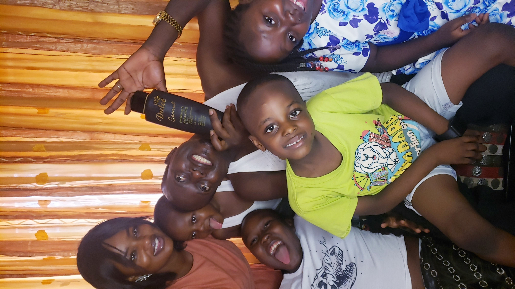 A group of six people are posing for a photo indoors. From left to right: a woman with short black hair wearing a reddish-brown shirt, an adult man holding a black bottle of “Dulce Carrot” lotion, two young children (one with their tongue out), a young child wearing a lime green shirt with a cartoon design, and a young woman in a blue and white floral shirt. All are smiling, and some are making silly faces. They are positioned at different angles, appearing to lean on and around each other. The background includes a golden-orange textured curtain or wall covering. The location is likely Sabon Tasha, Nigeria.