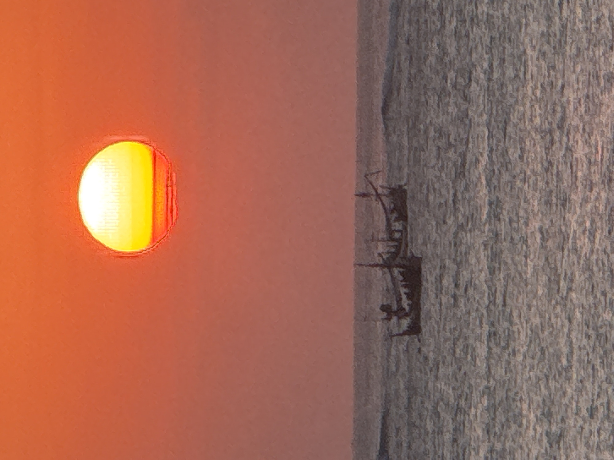 The image depicts a sunset over the ocean at Kuta, Indonesia. The sun, a bright orb of yellow and white, is low in the sky, casting a warm, orange glow across the scene. Two traditional Indonesian fishing boats, silhouetted against the horizon, are visible on the water. The sea is calm with gentle waves, reflecting the colors of the sunset. The sky is a gradient of orange and red, creating a serene and picturesque atmosphere. The boats are small, with distinctive triangular sails or roofs, and appear to be moving slowly. There are no people visible on the boats, suggesting they are either unattended or their occupants are not discernible from this distance. The overall impression is one of tranquility and natural beauty.