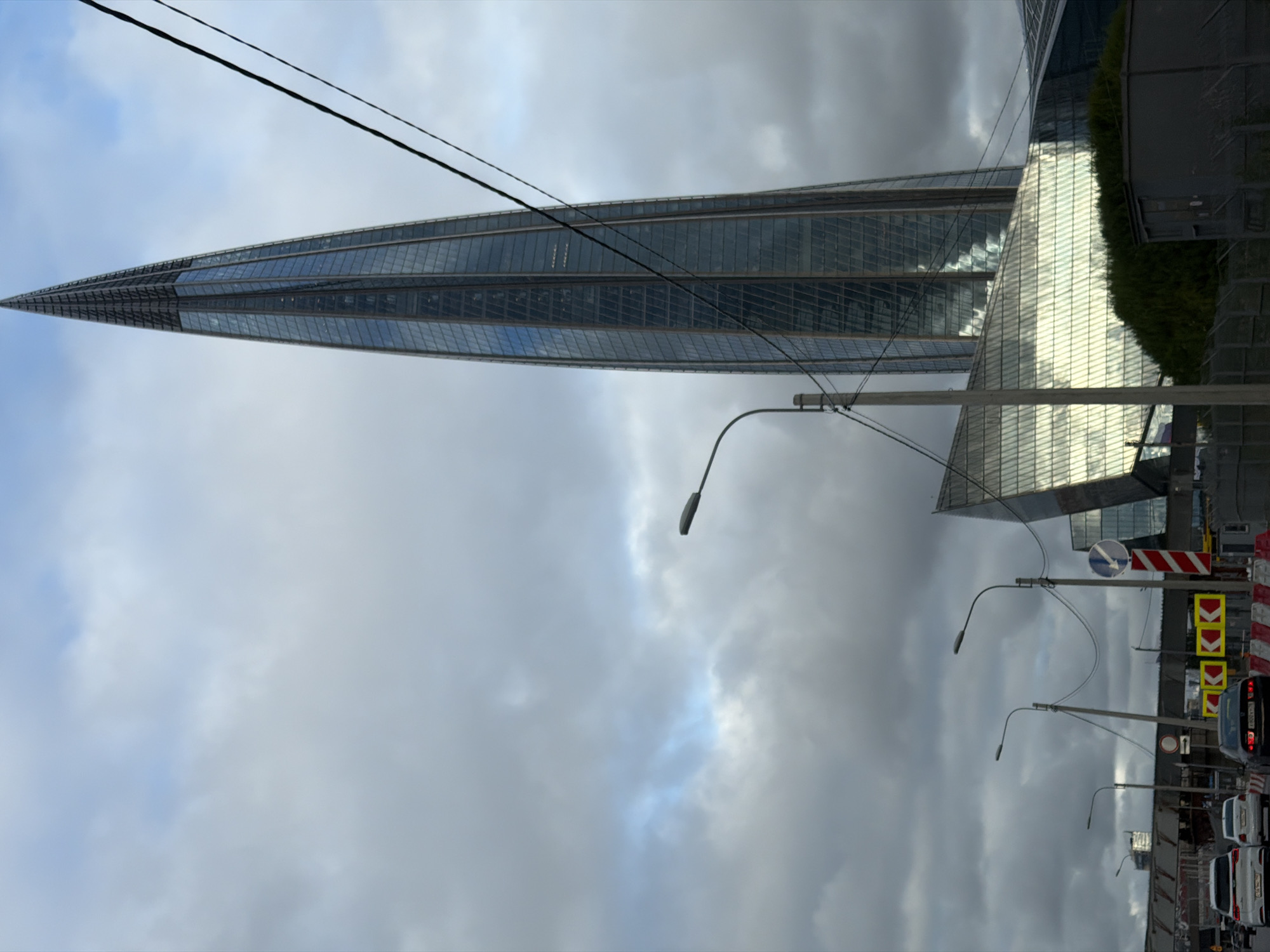 The image shows the Lakhta Center skyscraper in Saint Petersburg, Russia, on a cloudy day. The camera is positioned at street level, looking up towards the building. In the foreground, there is a road with several cars driving on it. Traffic lights and road signs are visible. The Lakhta Center dominates the upper right portion of the frame, its glass facade reflecting the cloudy sky. The lower part of the skyscraper is a wide, angled structure, also clad in glass. The sky is filled with gray, heavy clouds with some patches of lighter sky visible. The overall impression is of a modern urban environment under overcast weather conditions.