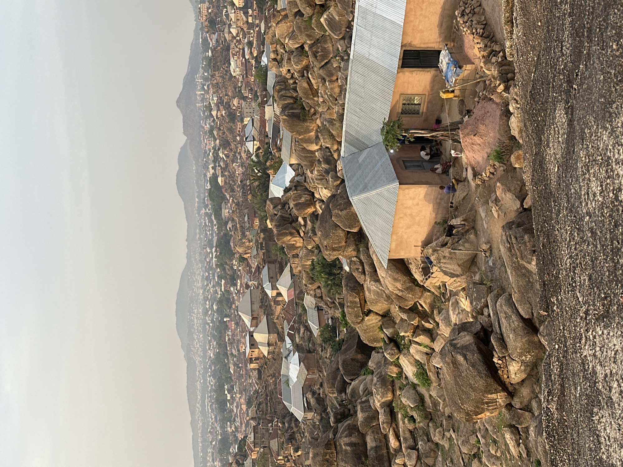 The image depicts a high-angle view of a residential area in Jos, Nigeria, characterized by buildings constructed amidst large rock formations. The houses have corrugated metal roofs and painted exteriors. Several individuals are present near one of the buildings, some are sitting and others are standing. The surrounding landscape is rugged, with exposed rock faces and scattered vegetation. A hazy atmosphere obscures the view of the distant hills.
