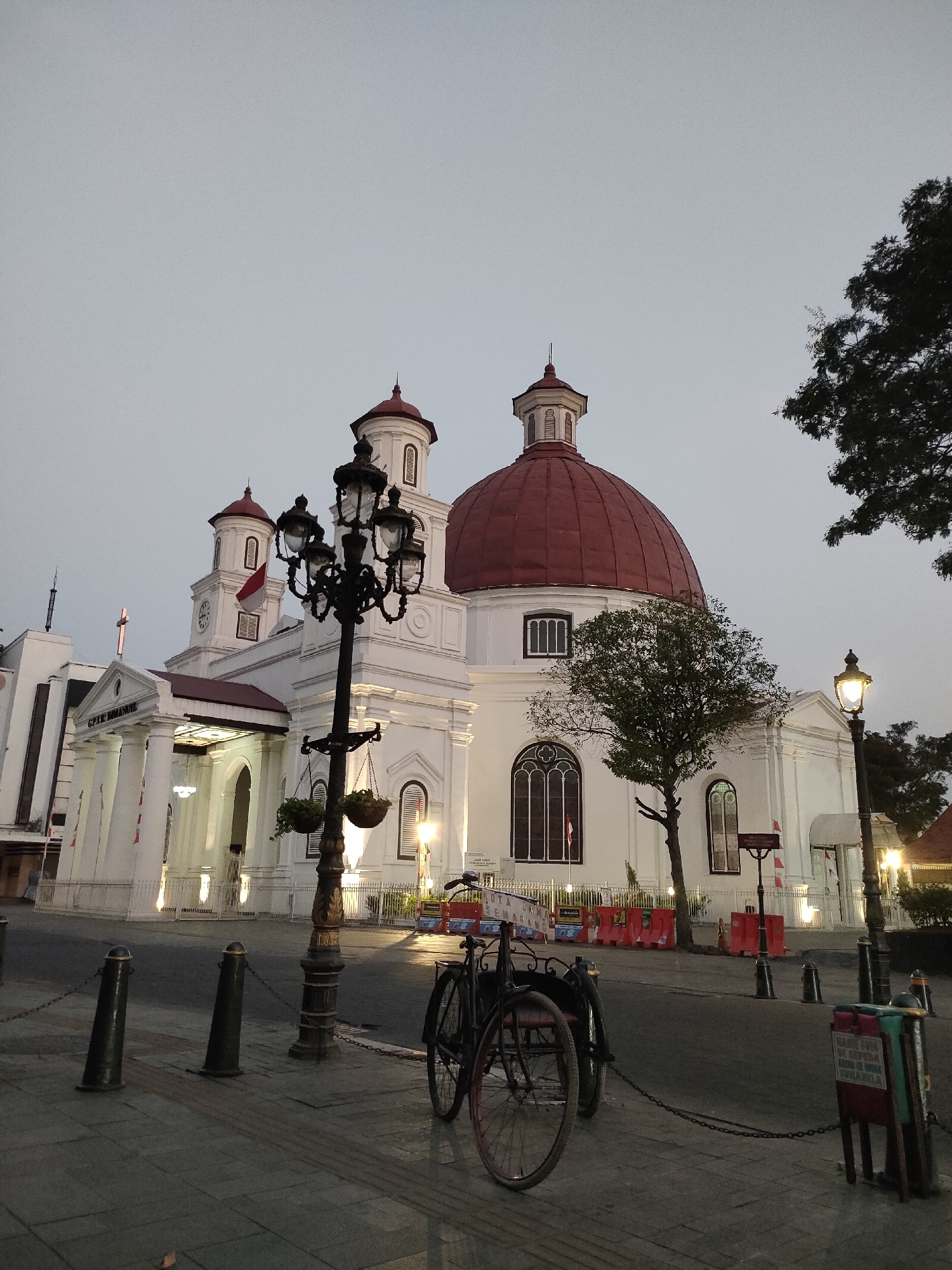 This is a ground-level, outdoor, medium shot of the Gereja Blenduk in Semarang, Indonesia. The church is a white, colonial-style building with a prominent red dome and two white towers topped with red roofs. The church's entrance features a portico with large white columns and an inscription above it. A large, ornate black lamppost stands in the foreground, with two hanging plants. In front of the church, there is a street with a sidewalk, bollards, chains, and a bicycle parked on the sidewalk. The sky is overcast and the lighting suggests it is either early morning or late evening. The scene is static, with no people visible.