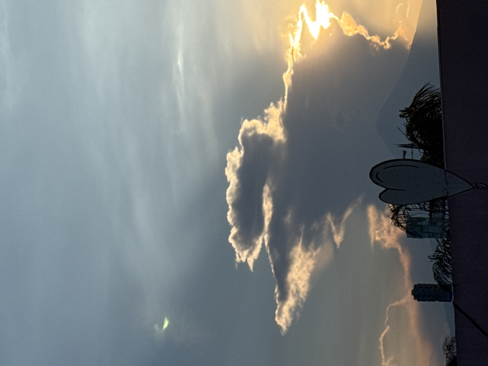 A daytime outdoor shot captures a cloudy sky in Puebla, Mexico. The sun shines brightly through a break in the clouds near the top right, with golden light illuminating the edges of the surrounding clouds. Below the clouds, the skyline of Puebla is visible, including buildings and palm trees. A satellite dish is prominently placed in the foreground. The overall scene is a blend of natural and urban elements.