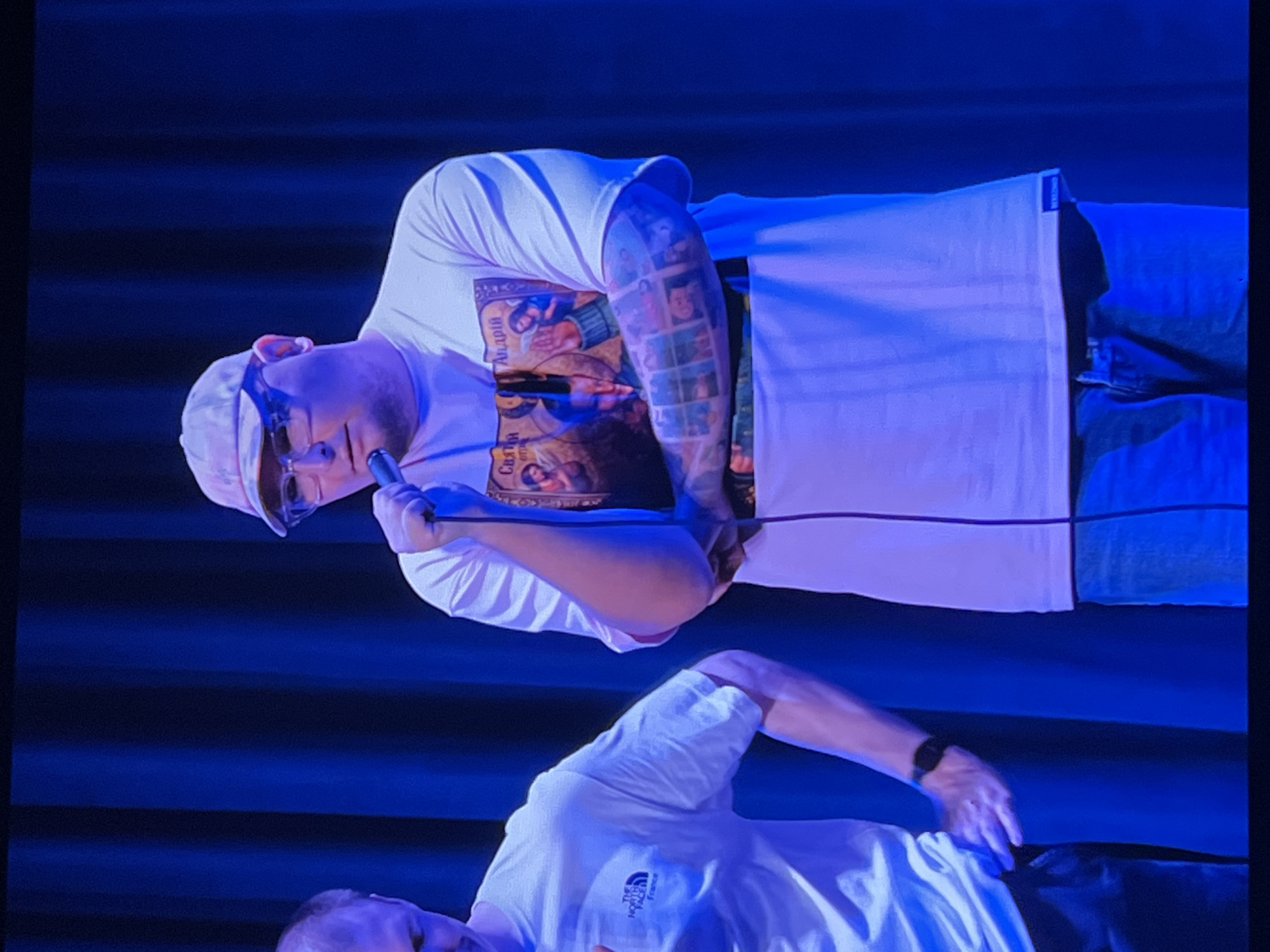 A man wearing a light-colored baseball cap, glasses, and a white t-shirt with a religious graphic and a tattooed arm is holding a microphone. He is standing in front of a dark blue curtain. Another man in a white t-shirt with "The North Face" logo is partially visible to the left, holding the first man's hand. The lighting is predominantly blue. The location is a stage in the city of Genemuiden, Netherlands.