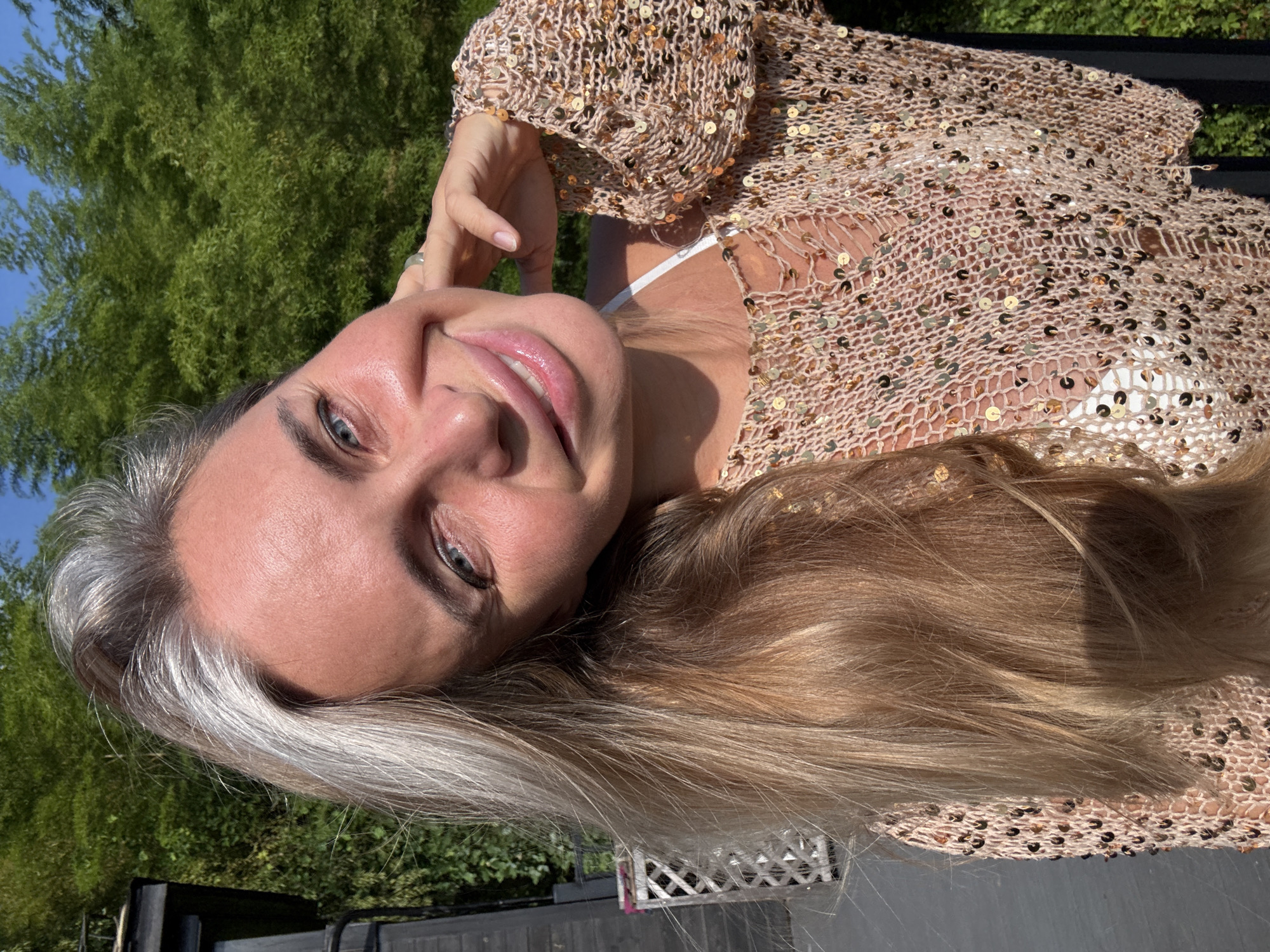 A selfie is captured from the shoulders up of a woman with blonde hair and striking blue eyes, her face turned towards the camera. She is smiling with her mouth slightly open, and her right hand is raised to her cheek, with a ring visible on her finger. The woman is wearing a light-colored, open-knit top adorned with sequins, revealing a white undergarment. Her hair is a mix of blonde and gray strands, falling loosely around her shoulders.
The background consists of lush green foliage, possibly bamboo, and a bright blue sky, suggesting an outdoor setting on a sunny day. To the left, a dark structure and a white lattice-like object are partially visible. The overall impression is one of a relaxed, sun-drenched moment. The image is devoid of any specific Wuxing District, China markers.