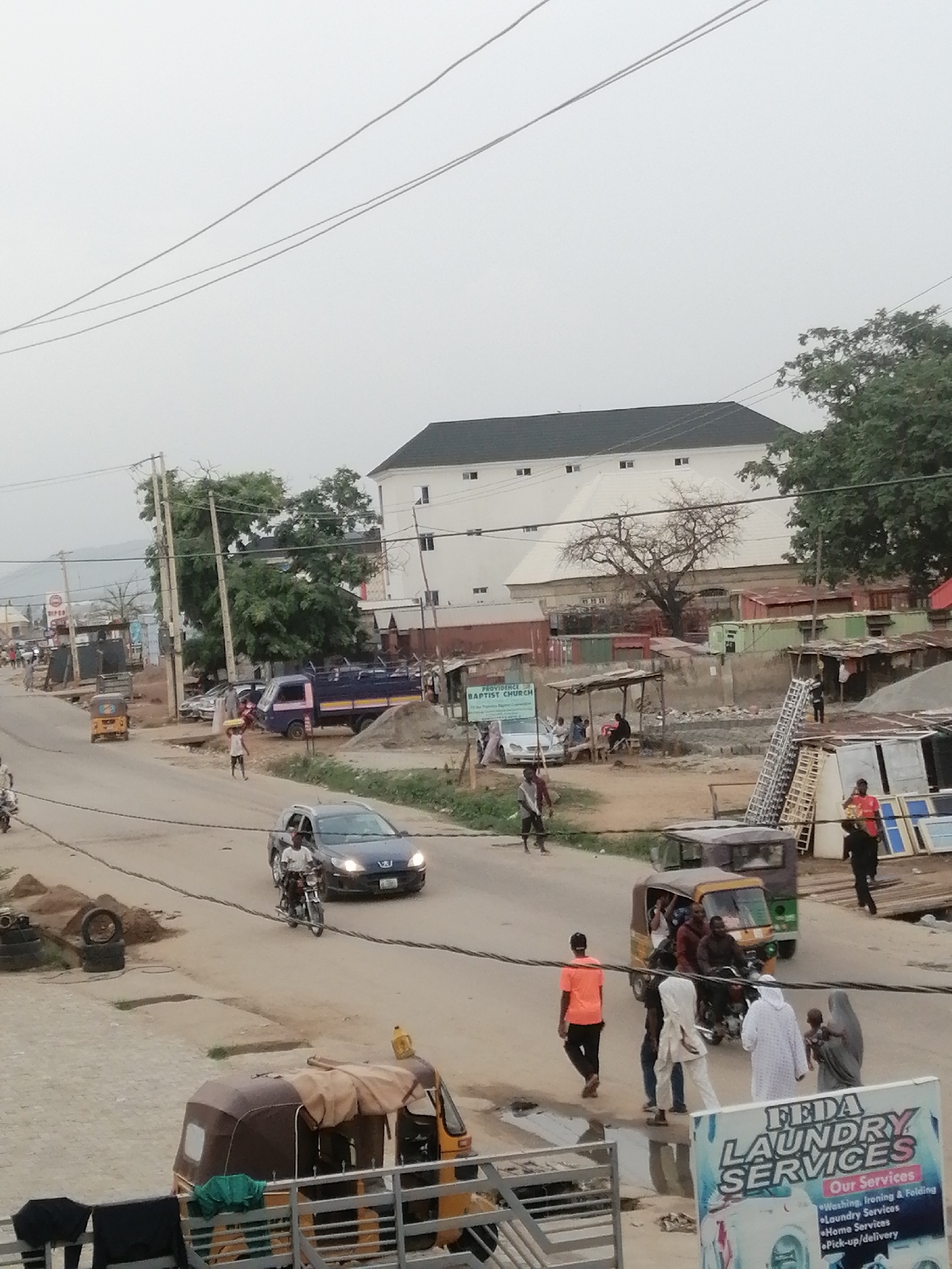 The image depicts a street scene in Sabojibi, Nigeria. The street is partially paved and lined with a mix of structures, including small businesses, residences, and a church. Several people are walking or gathered along the roadside, some in traditional attire. A few vehicles, including a car, motorcycles, and auto-rickshaws, are moving along the street. Power lines run overhead. A sign for a laundry service (FEDA Laundry Services) is visible in the foreground. In the background, a large, white building with a dark roof dominates the skyline. The overall atmosphere is one of a typical, busy day in a Nigerian urban setting.