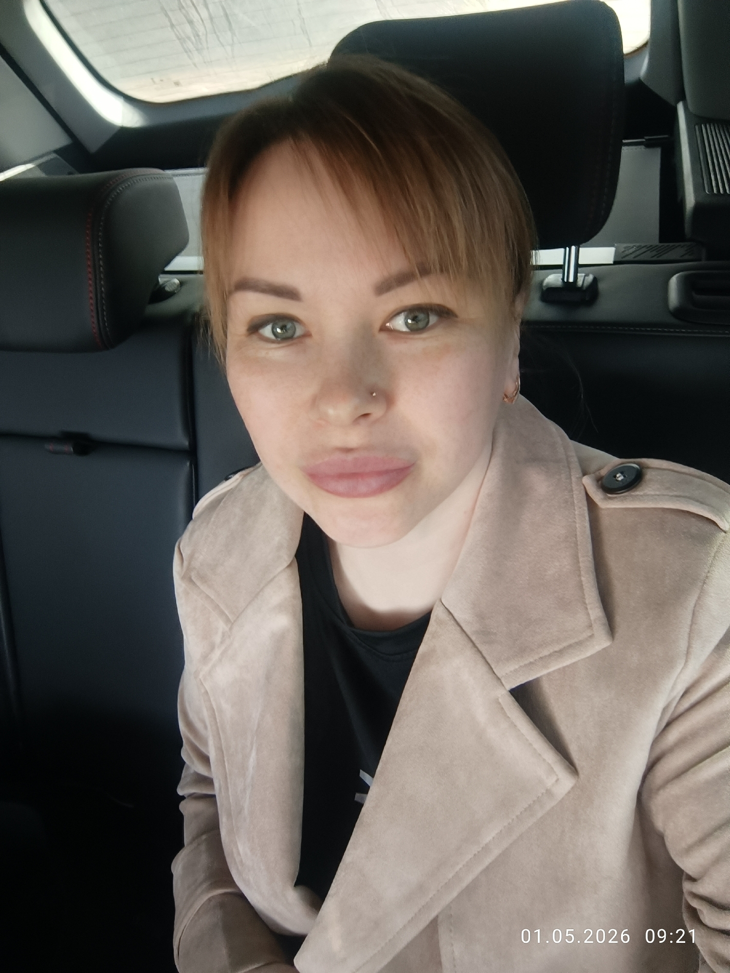 A woman is captured in a close-up selfie or portrait taken inside a car, likely in the back seat, in Chaikovsky, Russia. She looks directly at the camera with a neutral expression. Her short, reddish-brown hair features bangs across her forehead. She has green or hazel eyes, accentuated by eyeliner, and a small stud piercing on her left nostril. Her lips appear full and slightly parted. She is wearing a light beige or tan-colored jacket, possibly suede or faux suede, over a black top. The jacket has a wide collar and a visible black button on the right shoulder. A small hoop earring is visible in her right ear.
The background shows the interior of a car, including a black leather headrest with red stitching to her left. Behind her, a rear car window is visible, appearing somewhat streaky or dusty. The overall lighting suggests daytime.
In the bottom right corner of the image, a timestamp reads "01.05.2026 09:21".