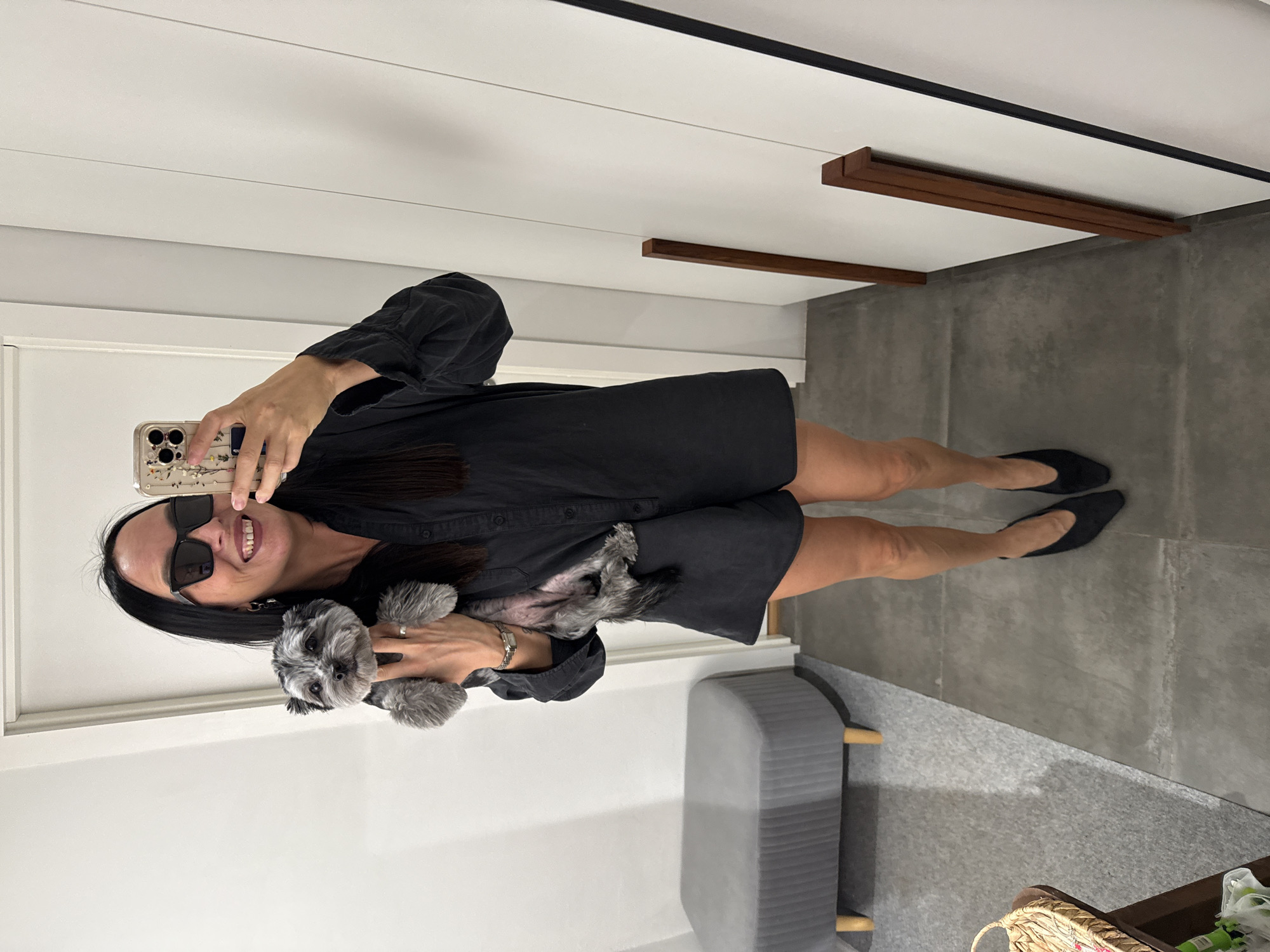 A woman with long dark hair, wearing large black sunglasses, a black collared shirt dress, and black pointed-toe heels, holds a small grey dog in her left arm. She is taking a mirror selfie with a smartphone. The background shows a modern interior with white walls, a grey upholstered bench, and a dark grey tiled floor. Two thin wooden planks are leaning against a white wall on the right side of the image. The scene is indoors, and there is no indication of the location being in Chaikovsky, Russia.