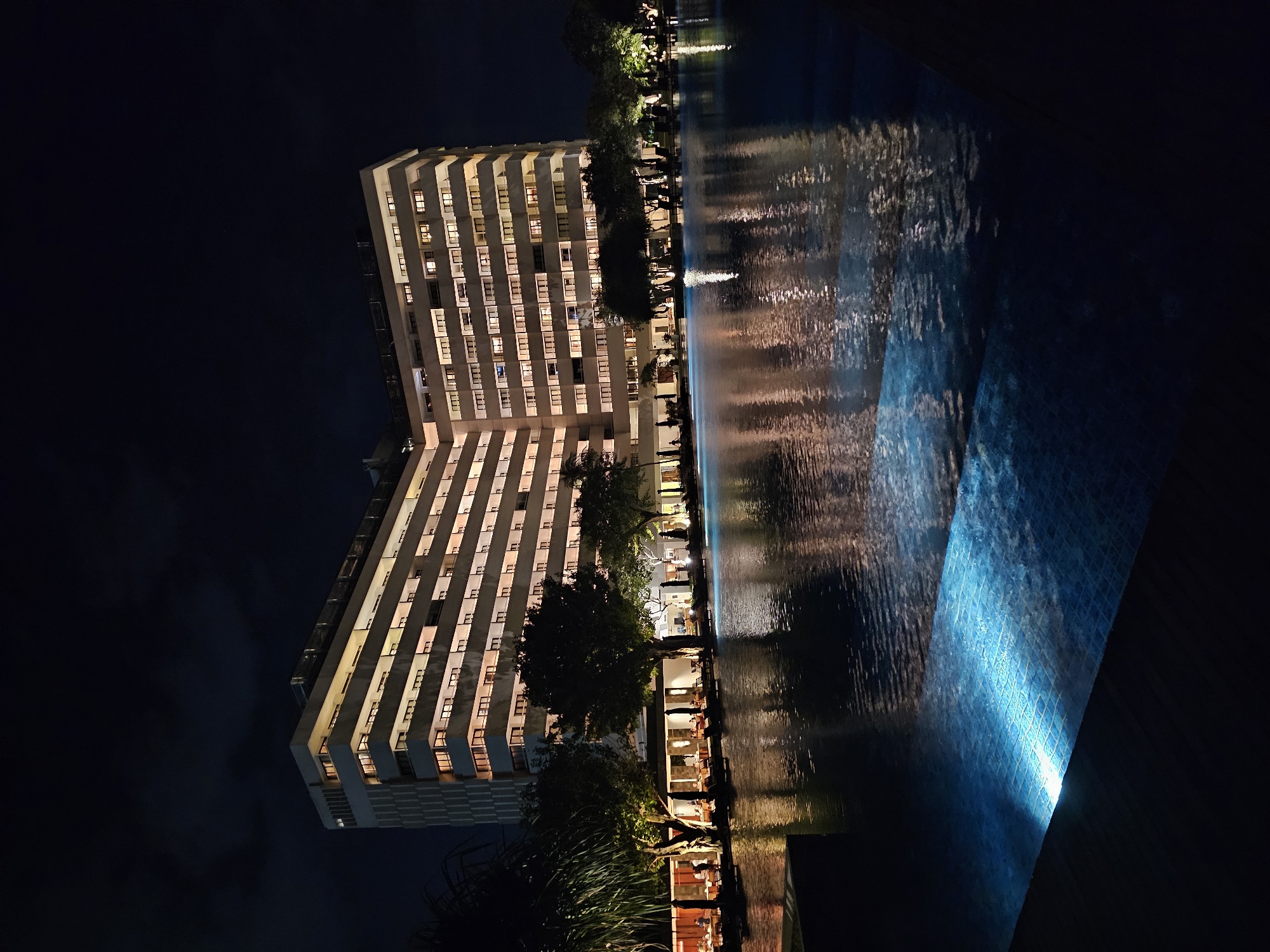 This is a night scene of a hotel in Denpasar, Indonesia, with a large swimming pool in the foreground. The hotel is a multi-story building with many windows, most of which are illuminated, suggesting it is occupied. The building's architecture is modern, with clean lines and balconies on each floor. The pool reflects the hotel's lights and the dark night sky. The water in the pool has an underwater light, casting a bright blue glow on the tiled bottom in the foreground. Trees and some outdoor seating areas are visible between the hotel and the pool. There are a few people visible in the seating area, and some are standing and walking. The overall impression is one of a well-lit and inviting resort at night.