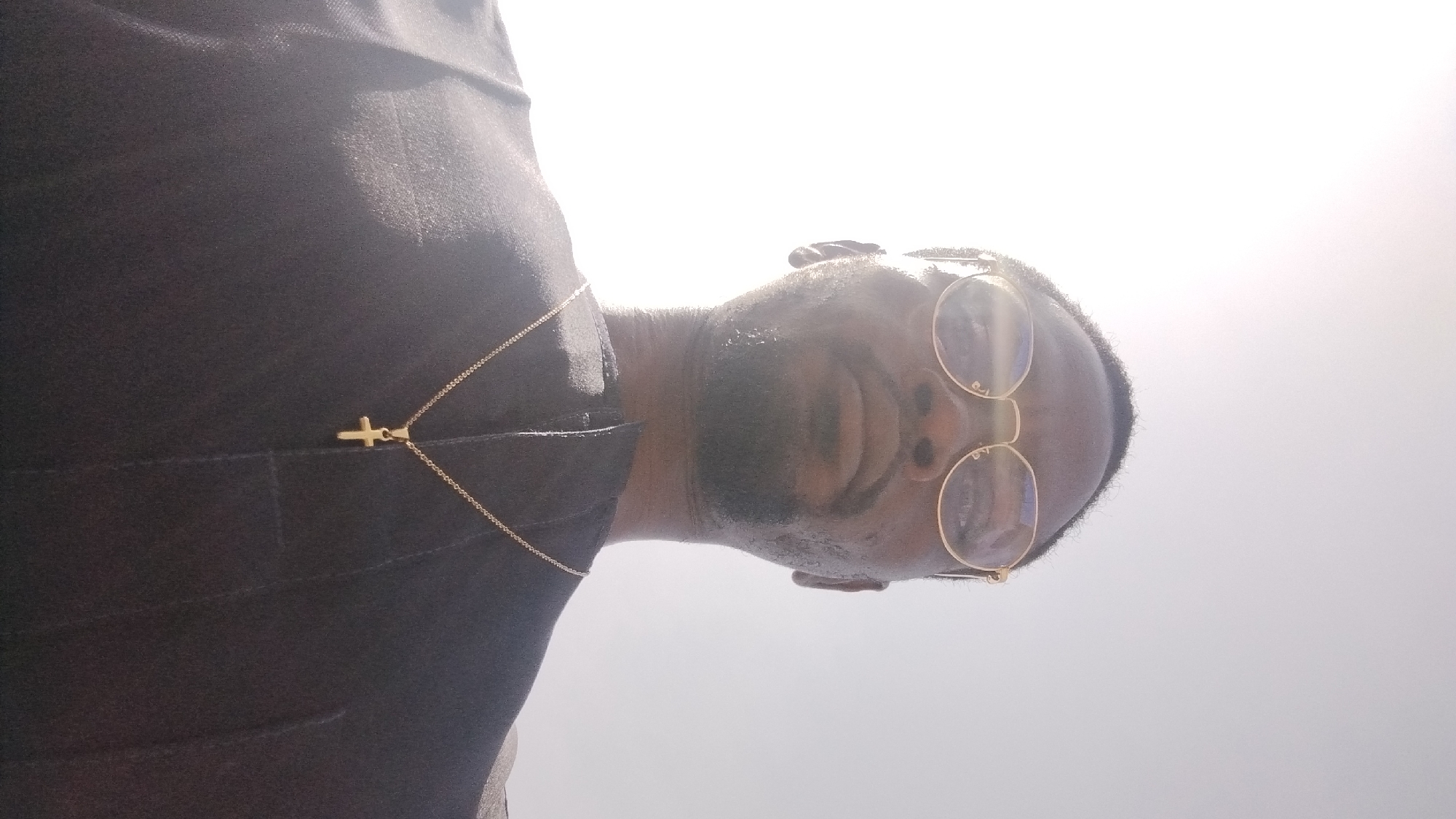 A low-angle, close-up shot shows a man wearing a dark garment and gold-rimmed glasses, looking upwards. A gold cross necklace hangs on his chest. The background is a bright, blown-out sky, possibly indicating an outdoor setting. The image's lighting and composition suggest it may have been taken in Jos, Nigeria, given its bright sunlight and the subject's attire, which could be traditional or formal wear.