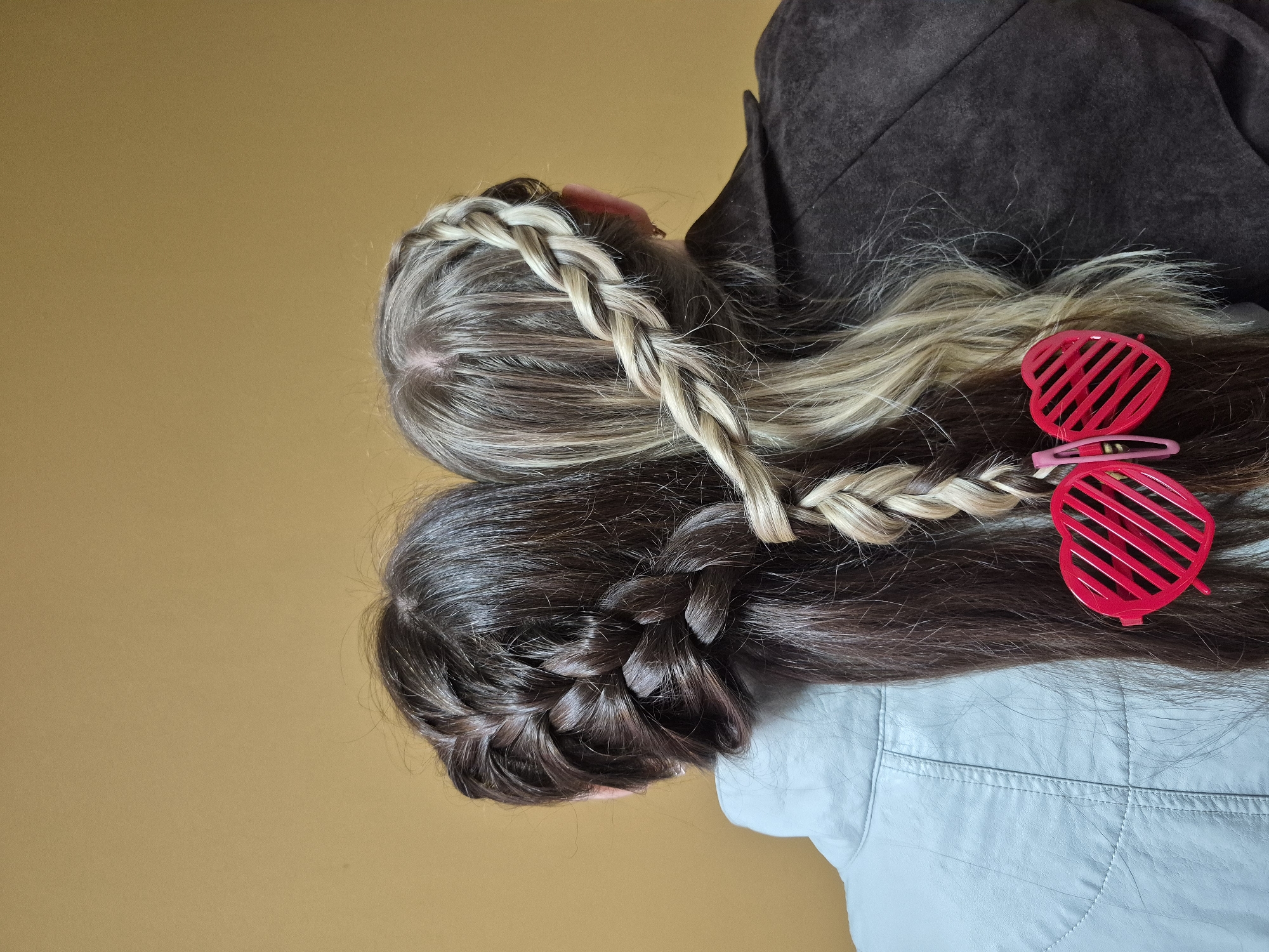 This vertically oriented close-up image captures the backs of two individuals with distinct hairstyles, set against a plain mustard yellow background. The scene is located in Chaikovsky, Russia.
On the left, an individual with multi-toned light brown and blonde hair is visible, wearing a dark brown jacket that appears to have a suede or textured finish. Their hair features a prominent light-colored braid that originates from their head and gracefully crosses over, intertwining with the hair of the second individual.
On the right, a second individual with long, dark brown hair is present. Their hair is styled with a French braid that extends down the back of their head. They are wearing a light blue or grey jacket, characterized by visible stitching details across the back. Resting on their dark hair, partially obscured, are bright red heart-shaped shutter shades and a small pink hair clip.
The intertwined braid between the two individuals suggests a playful connection or a shared artistic hairstyle. The plain background indicates an indoor setting, possibly a studio or a simple wall. No faces are visible, so emotions cannot be discerned, nor can specific ages. The time of day and weather are not determinable from the image. There is no visible text.