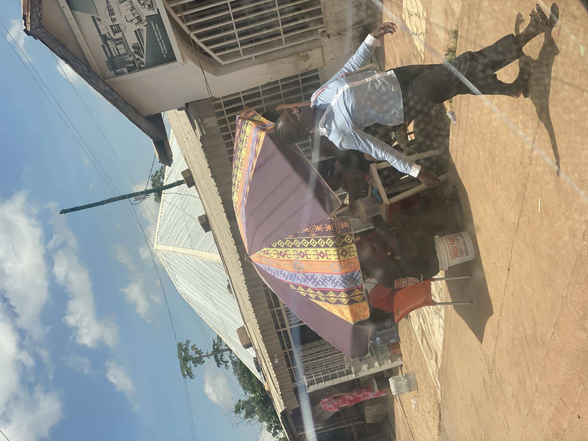 The image depicts a street scene in Gura Top, Nigeria, under a partly cloudy sky. A woman in a blue and white striped shirt and dark pants is walking across a dusty, paved area. She is carrying a light-colored bag over her right shoulder and is looking to her right. To her left, an umbrella with colorful geometric patterns shades a small seating area where other individuals are visible. Behind the umbrella, there are several structures, including a building with a metal roof and a building with a sign that appears to be advertising architectural services. In the background, trees and utility poles are present. The ground is a mixture of dirt and pavement, with some debris scattered around.