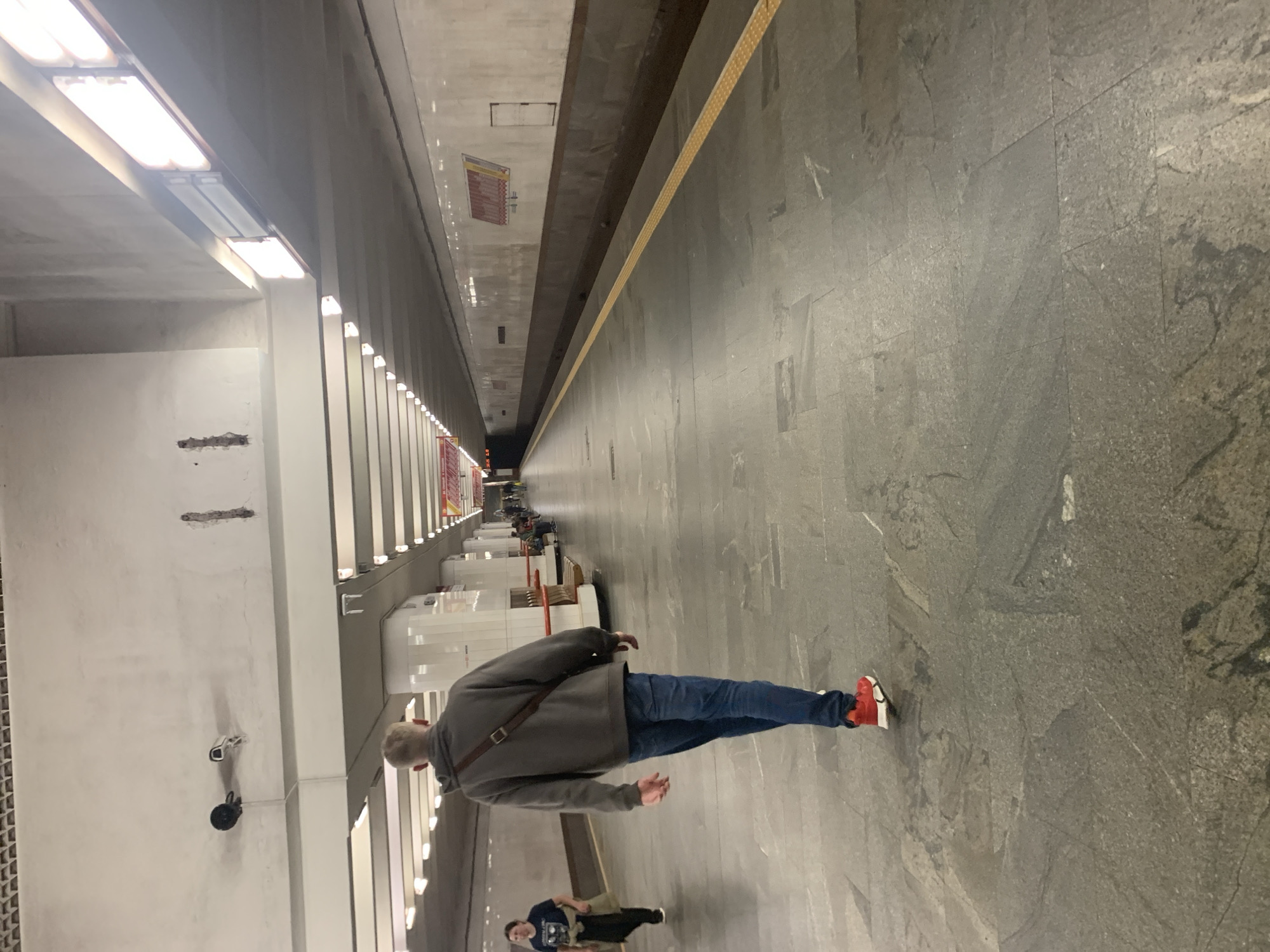 A man is walking away from the camera down a subway platform in Minsk, Belarus. He is wearing a grey hooded sweatshirt, blue jeans, and red sneakers. He has a brown bag slung over his left shoulder. Another person, a woman, is walking in the opposite direction on the left side of the platform. The platform is paved with large grey stone tiles. A yellow tactile warning strip runs along the edge of the platform. The ceiling of the station is white, with rows of fluorescent lights. Two security cameras are visible on the ceiling. The walls of the station are tiled in a lighter color with a darker band across the middle. There are benches and columns along the platform. Signs are visible on the walls in the distance.